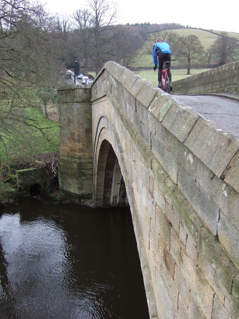 A stone arch bridge over an English river, its wedge-shaped stones curving over the water while a cyclist crosses above