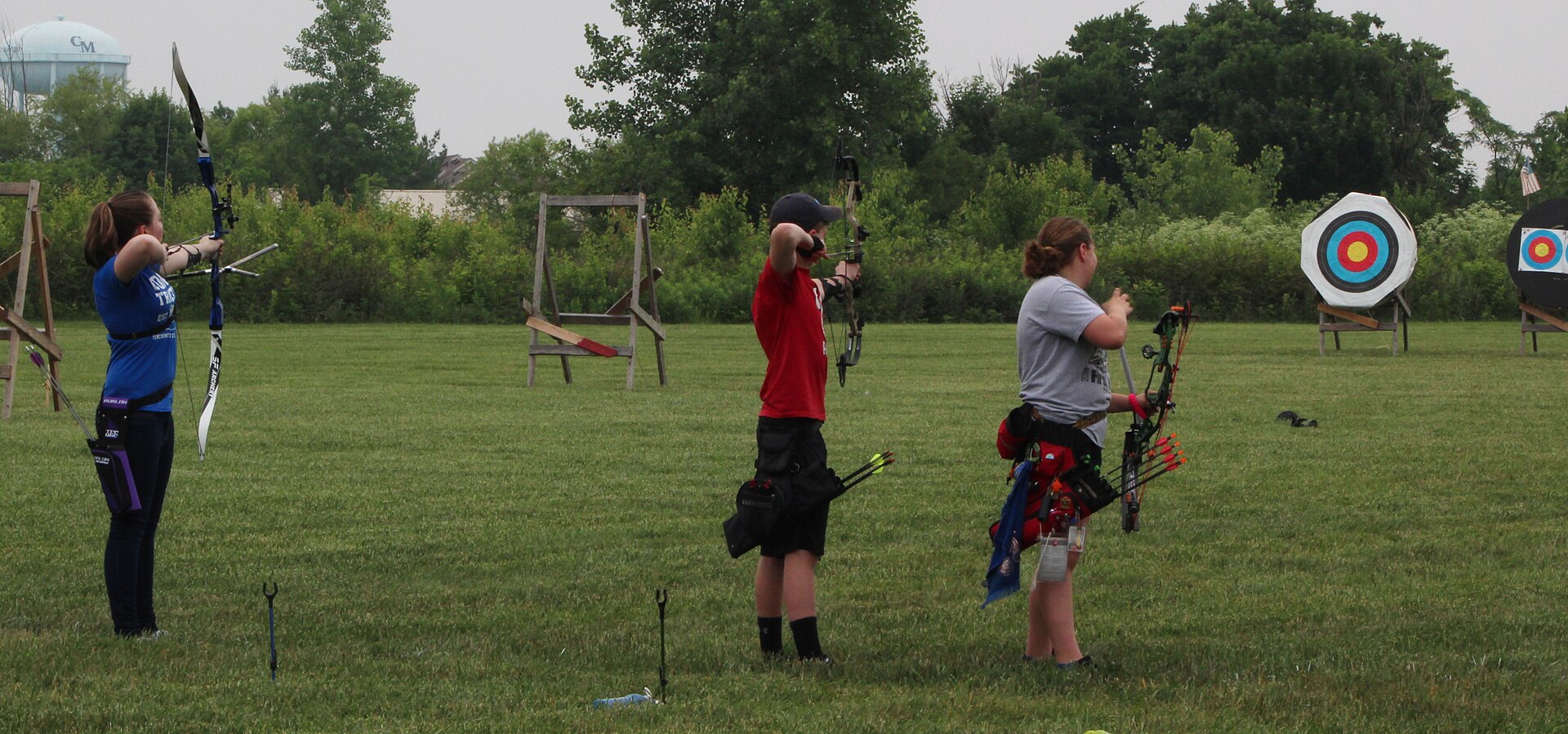 Three archers standing on a grass field with bows drawn, aiming at round target faces some distance away.
