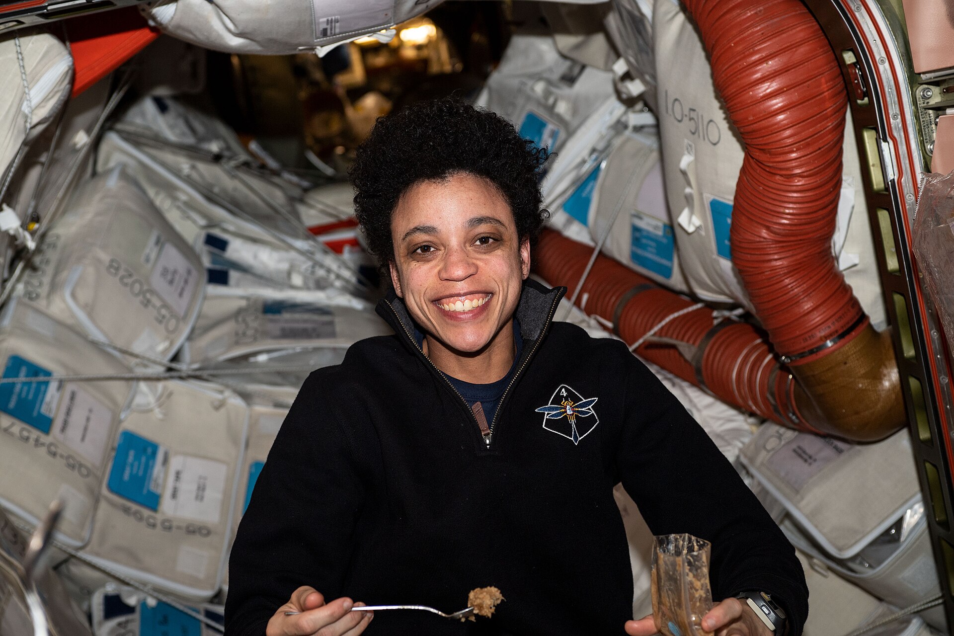 An astronaut aboard the space station smiling with a spoonful of food and a sealed drink pouch