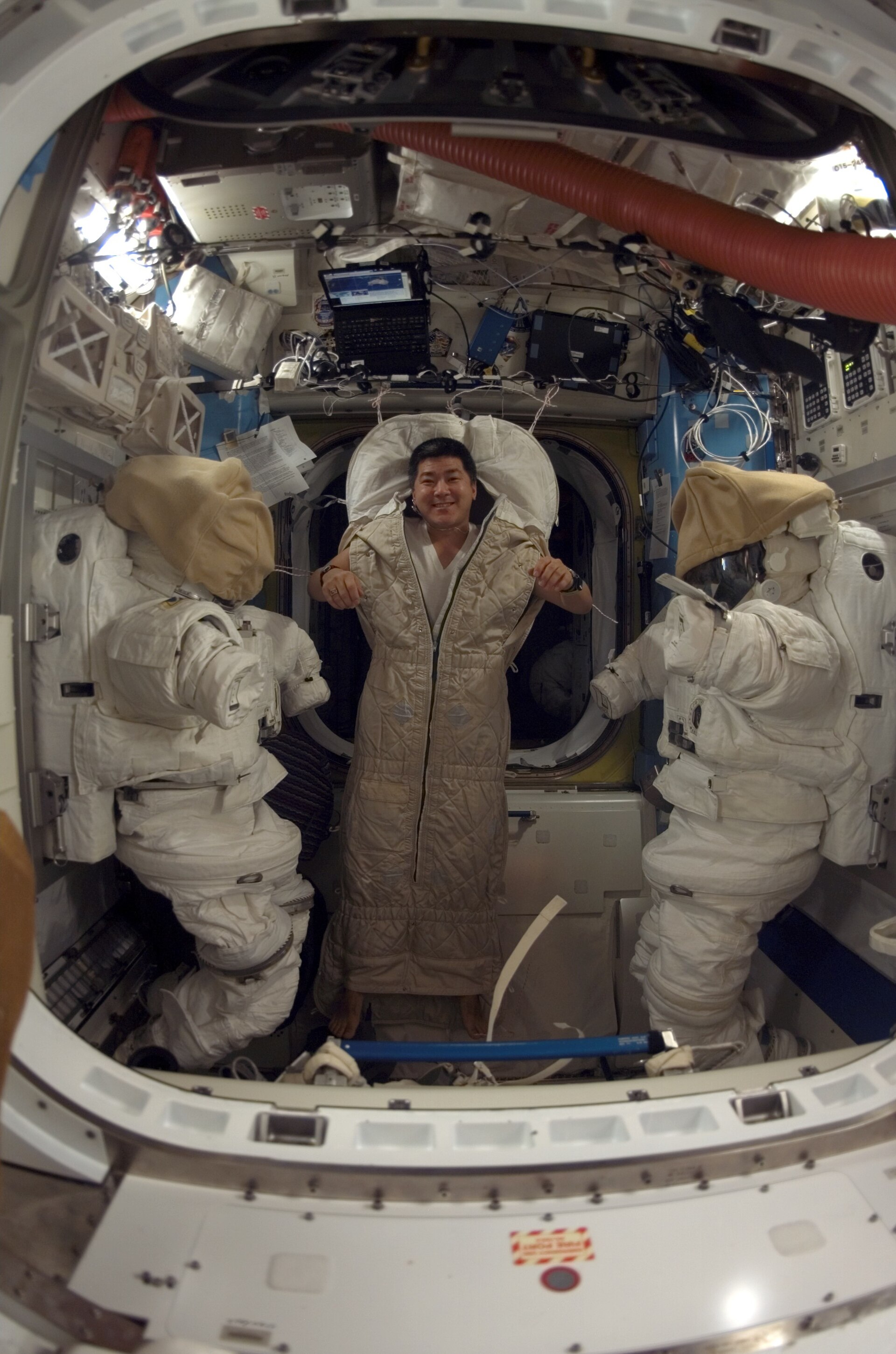 An astronaut floating inside a padded sleeping bag on the space station, flanked by two spacesuits