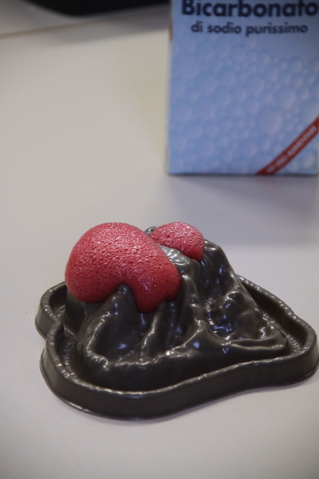 A model volcano erupting with red foam from a baking soda and vinegar reaction, a box of sodium bicarbonate behind it.