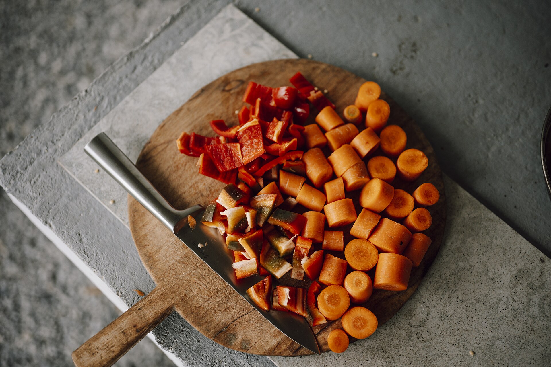 Chopped carrots and red peppers piled on a round wooden board with a large knife resting beside them.