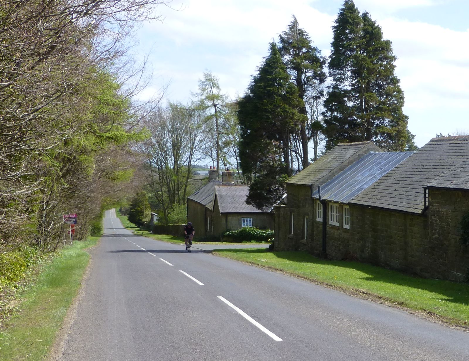A lone cyclist grinding up a long climb on a quiet country lane past stone cottages, the kind of unaccustomed effort that leaves legs aching the next day