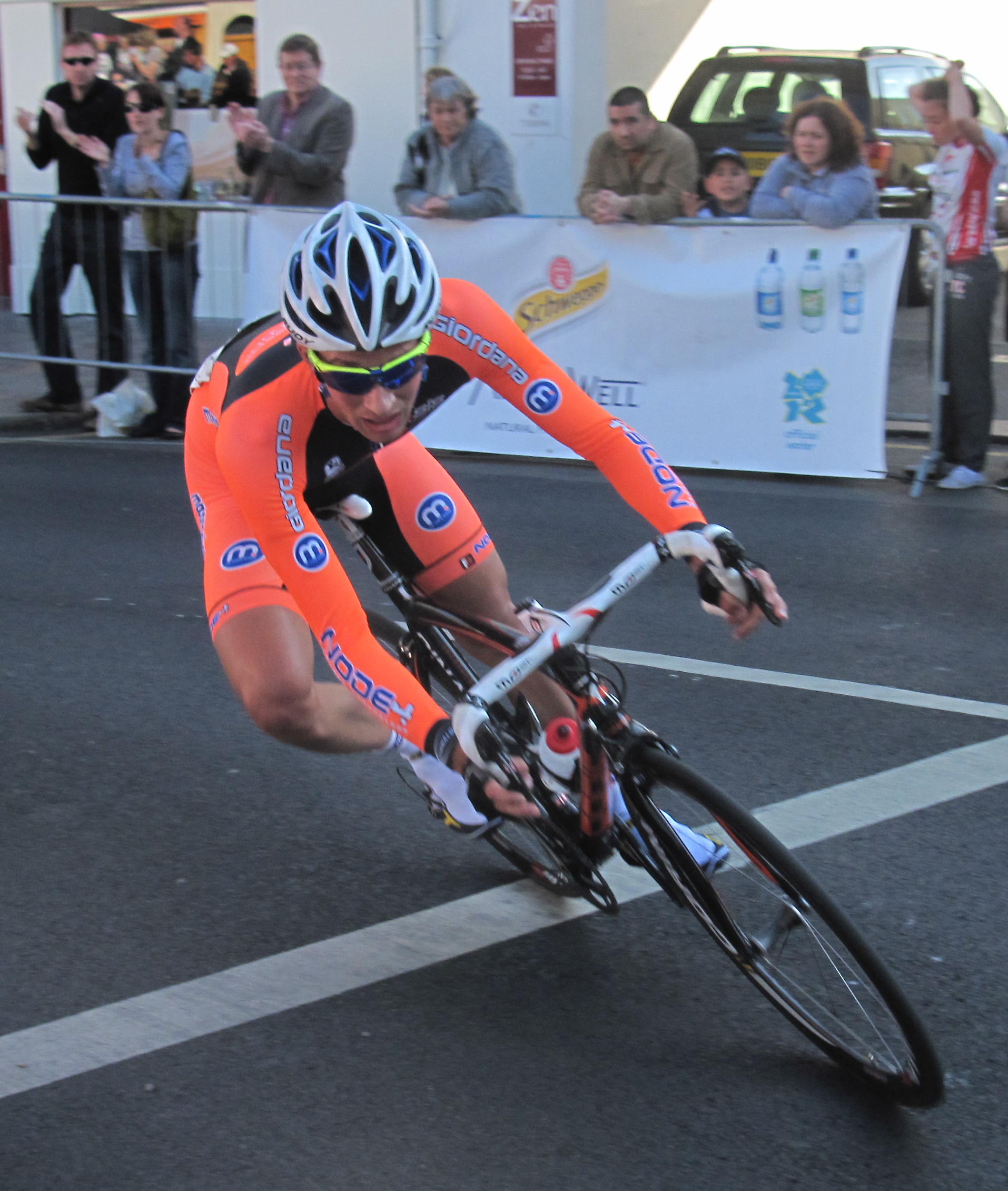 A racing cyclist leaned steeply over into a corner on a town-centre road, bike and rider tilted well in towards the inside of the bend.
