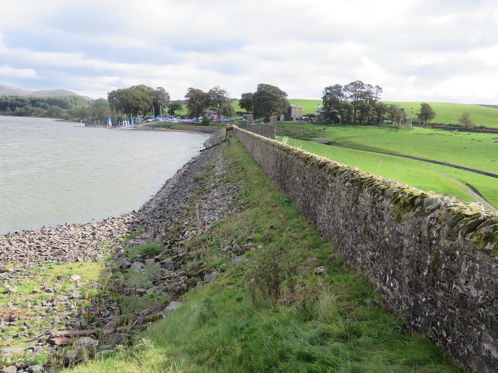 A stone dam holding back a reservoir, its wall broad at the base and topped by a stone parapet.