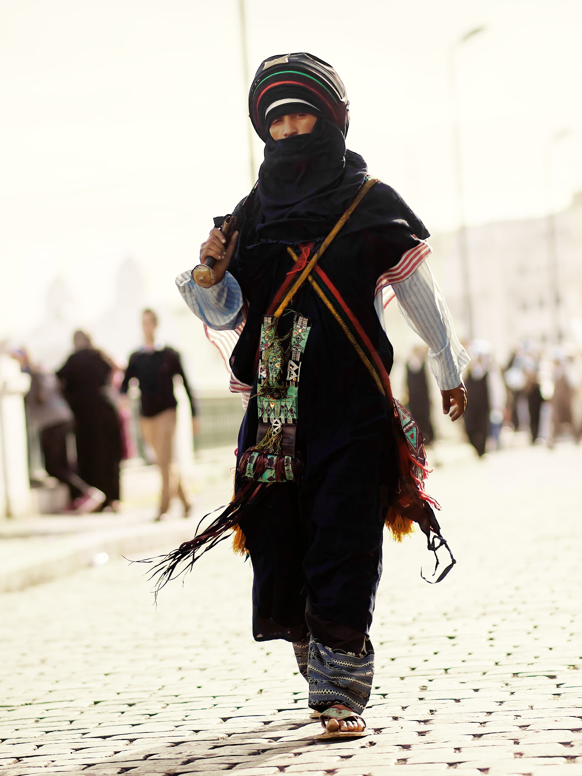 A Tuareg man walking in bright sunshine wearing loose flowing dark robes and a wrapped head covering.