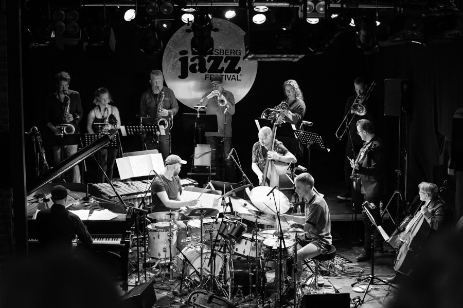 A jazz orchestra on stage with instruments of every size together: a tall double bass and cello beside small and large saxophones, trumpet and tuba