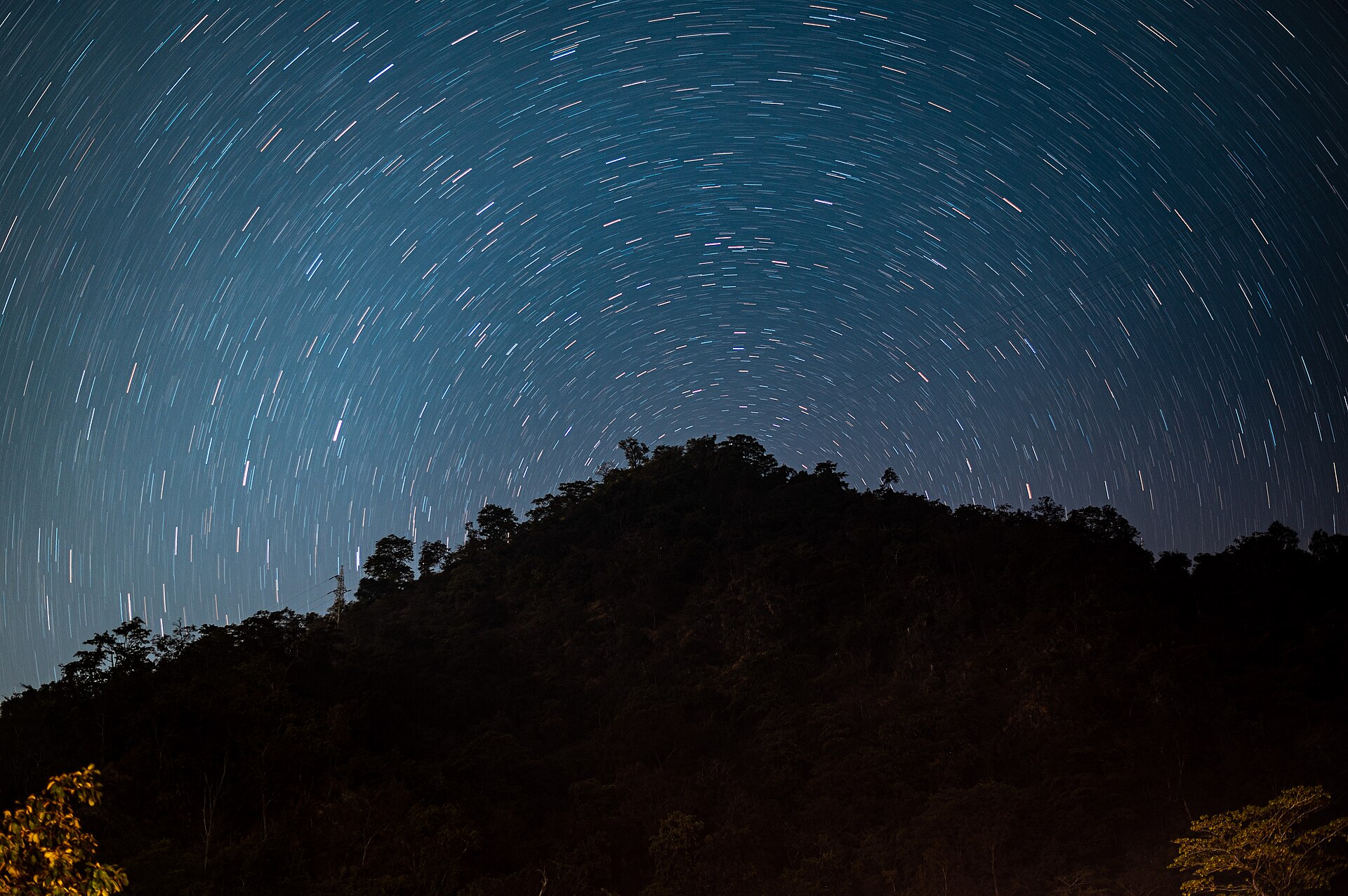 A long-exposure night photograph in which the stars have drawn concentric arcs across the sky above a dark wooded hill, tracing the Earth's turn.