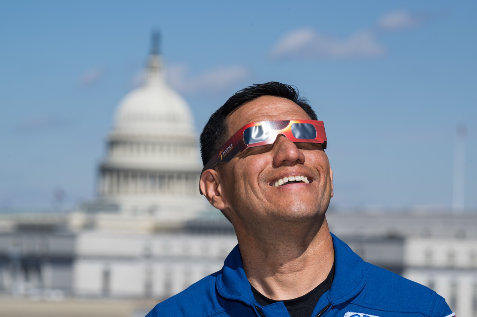 A smiling man wearing special cardboard eclipse glasses looks safely up toward the Sun.