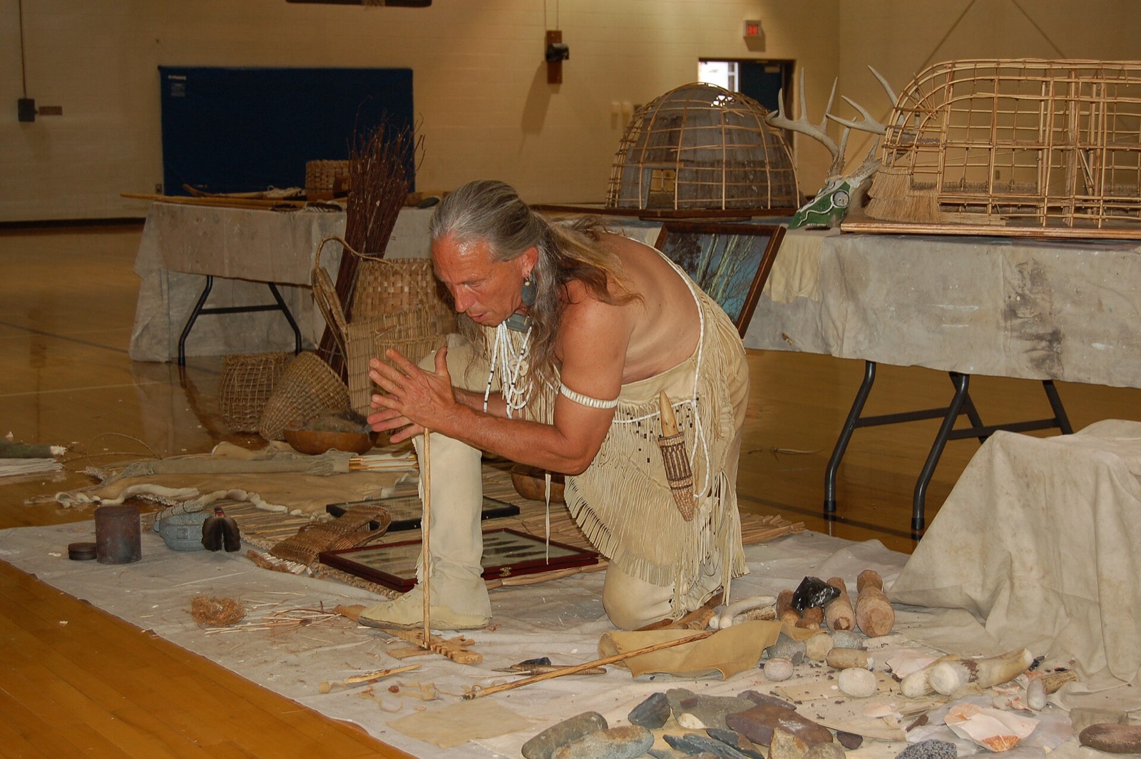 A person spinning a wooden stick between their hands against a board to make heat by friction and start a fire, the way people did long ago.
