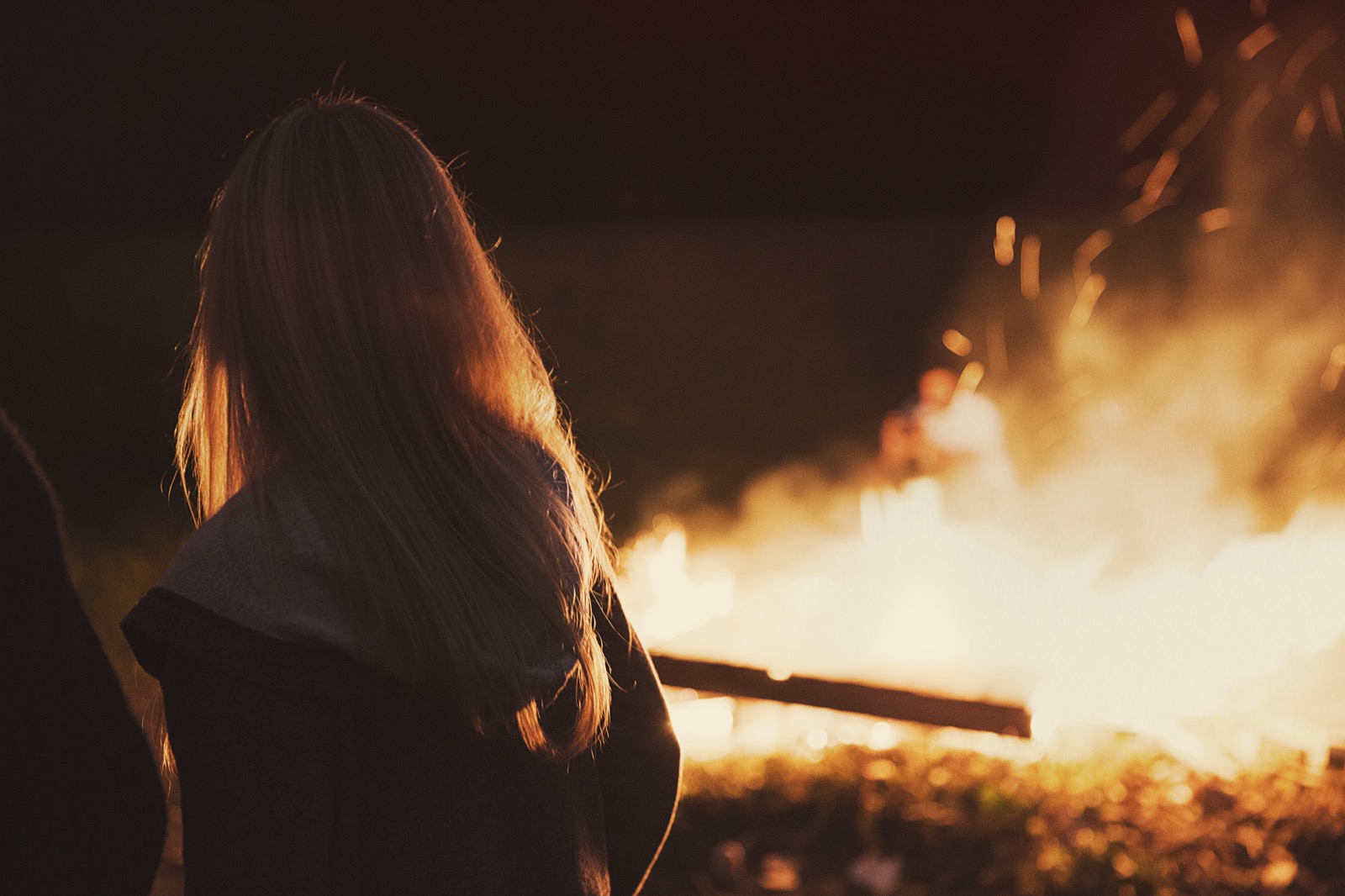 A person watching a glowing campfire from a safe distance at dusk, flames and warm light against the dark.