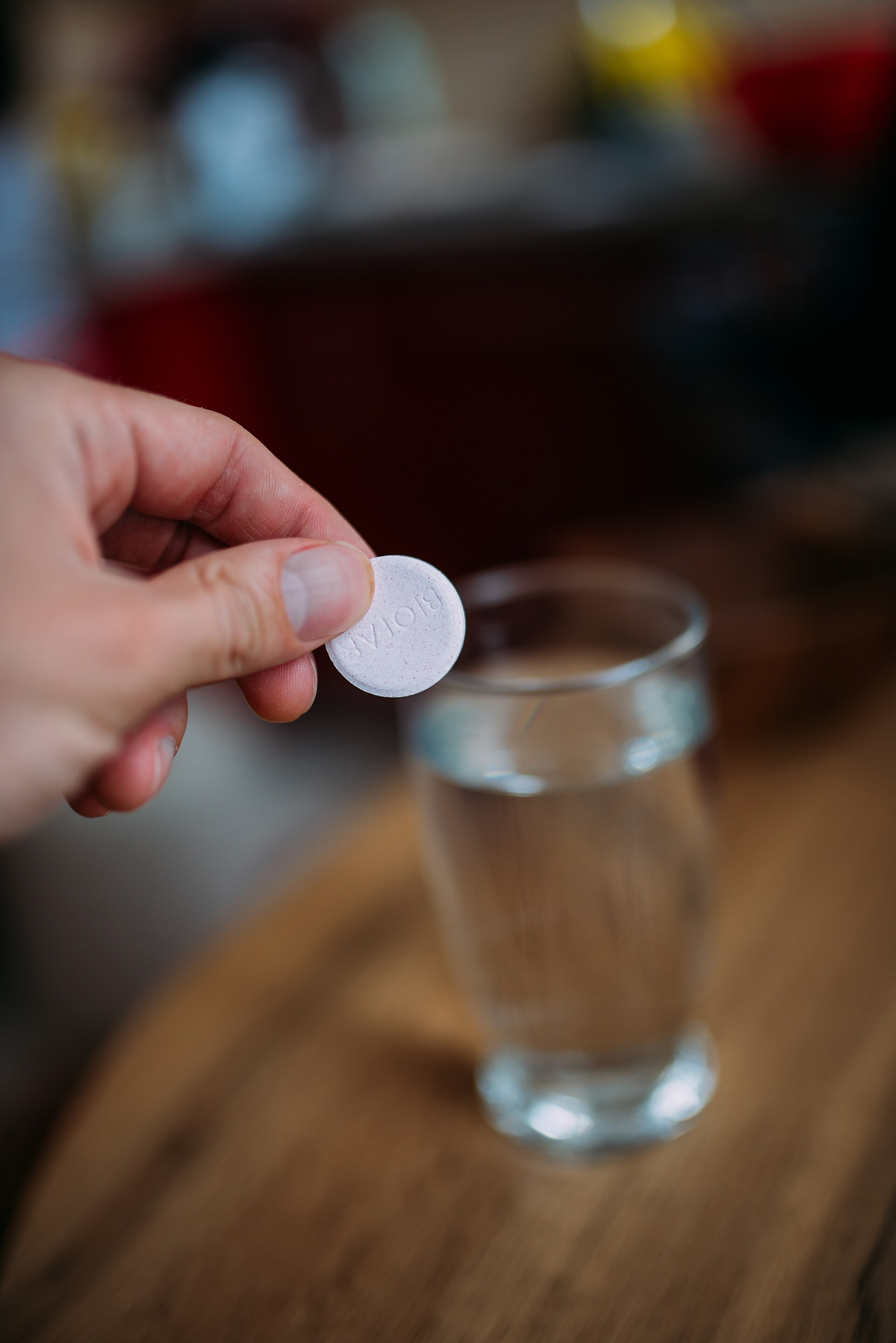 A hand holding a white effervescent tablet above a glass of clear water, about to drop it in.