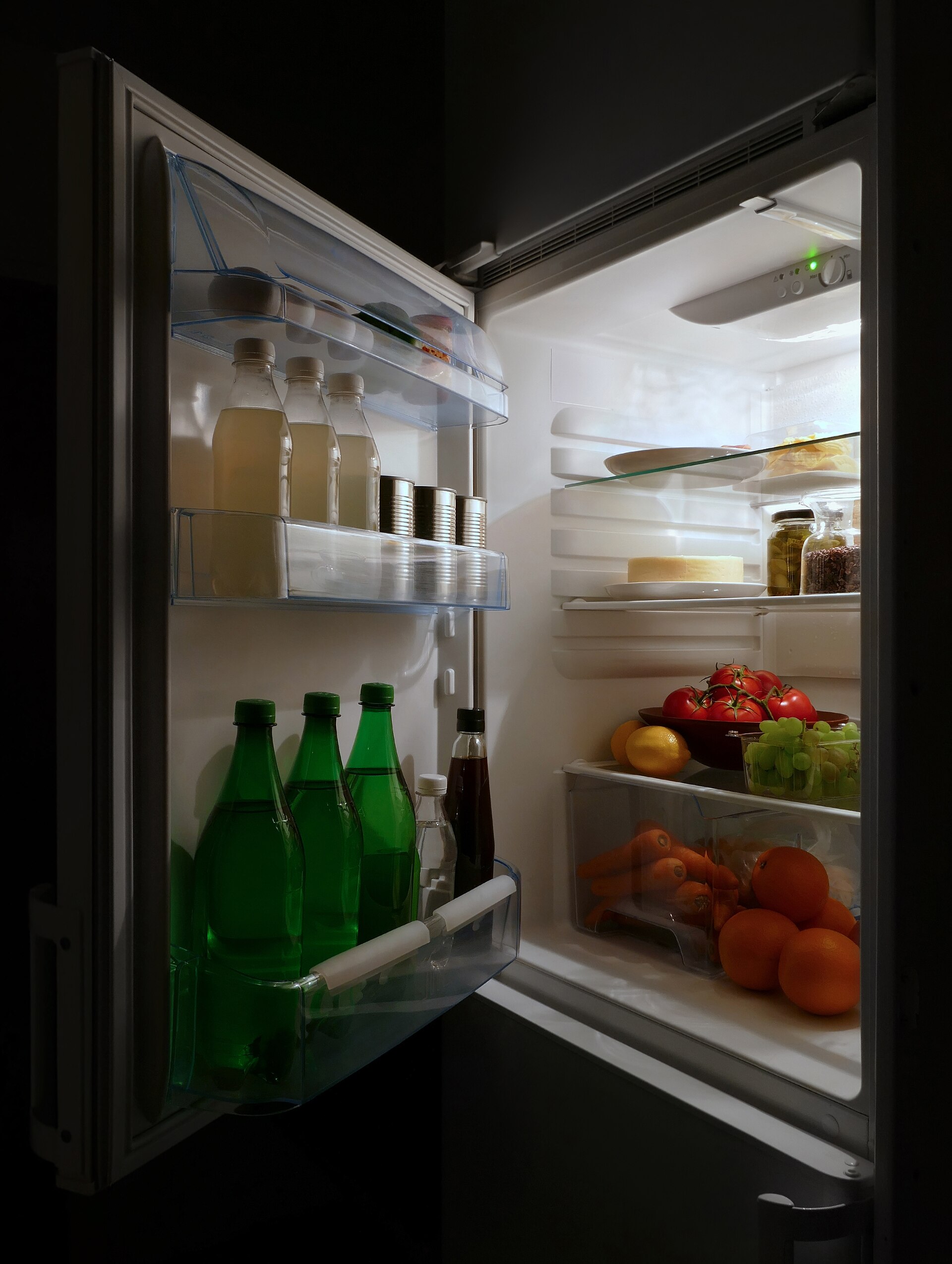 An open fridge lighting up a dark kitchen, its shelves full of milk, cheese, tomatoes and oranges