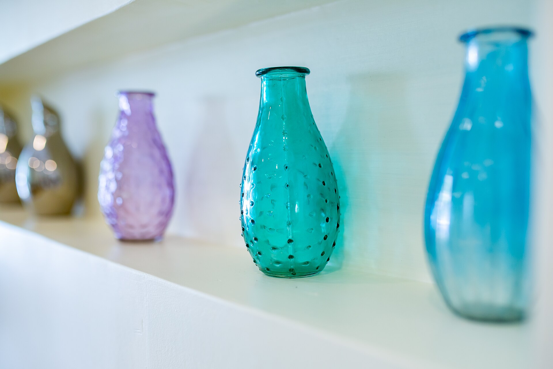 Coloured glass bottles in a row on a white shelf, each with a narrow neck opening into a rounded body of air