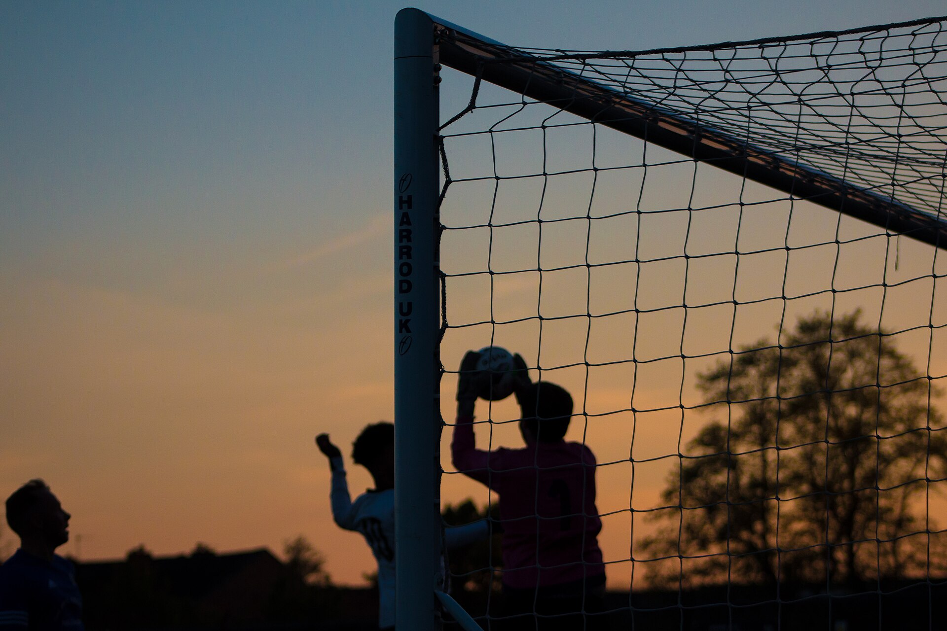 A goalkeeper silhouetted against an orange sunset sky, catching a football with both hands in front of the goal net.