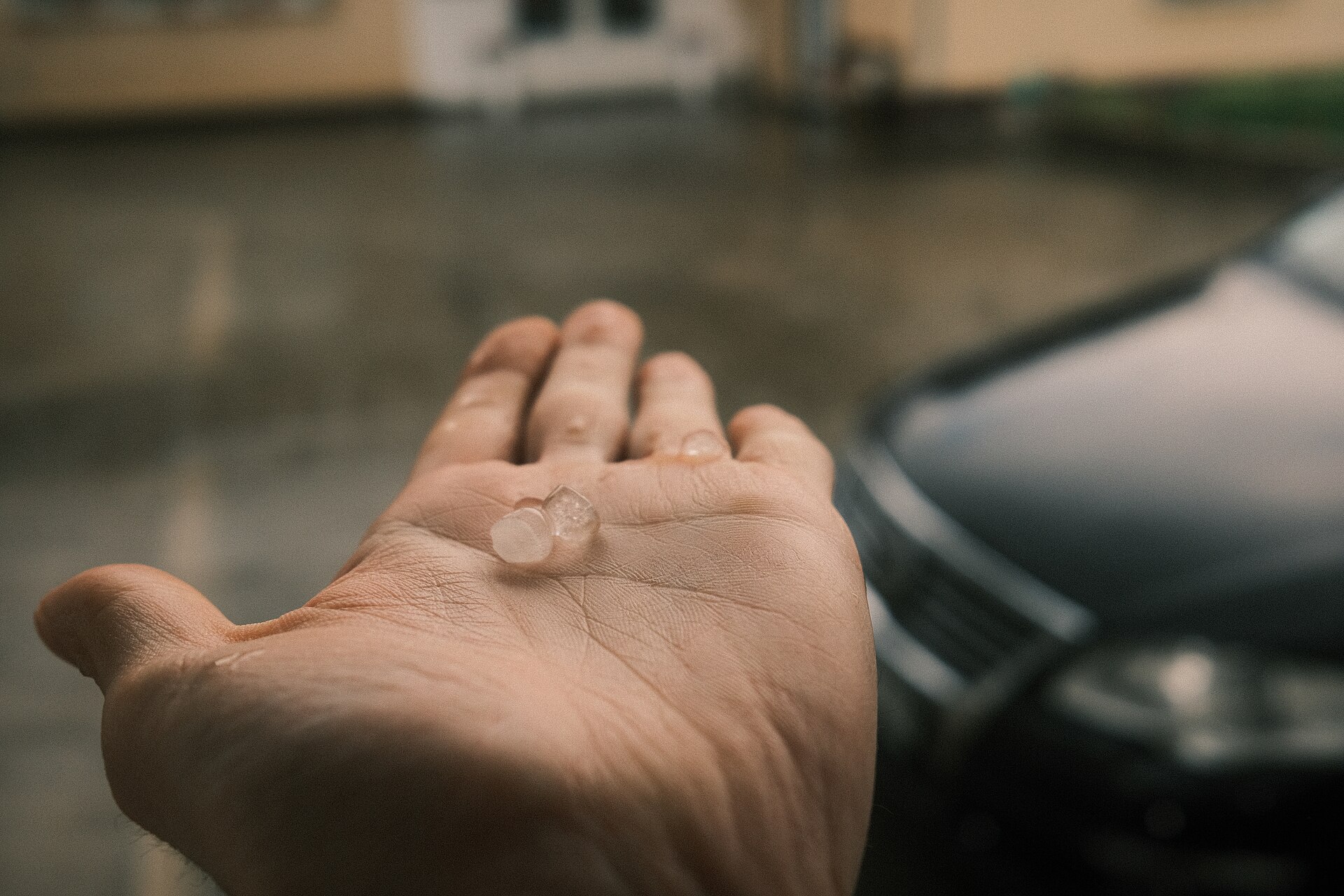 An open palm holding two small clear lumps of hail, with wet ground and a parked car blurred behind.