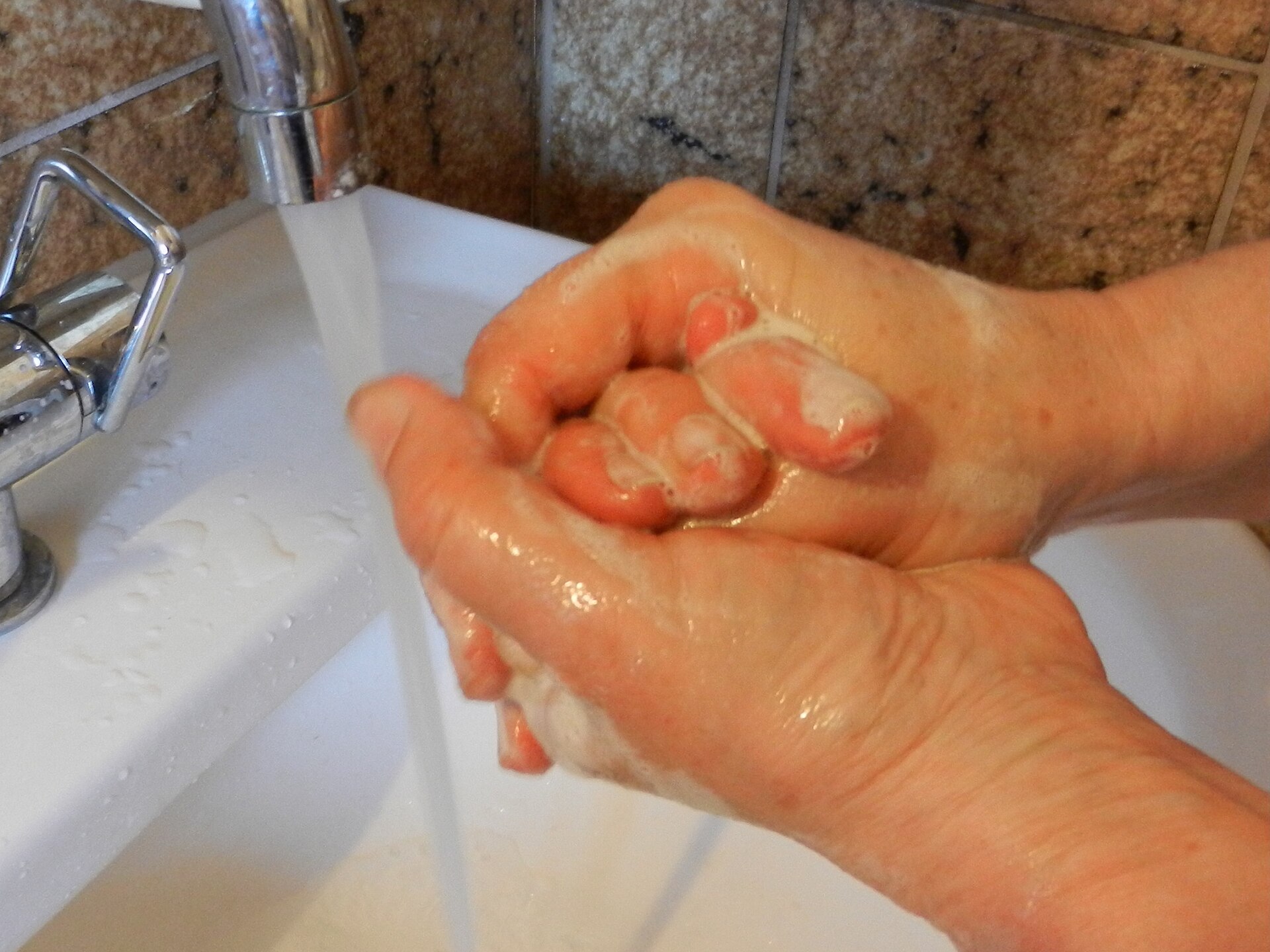 A pair of hands being lathered with soap under a running tap, fingers interlocked to work the suds between them