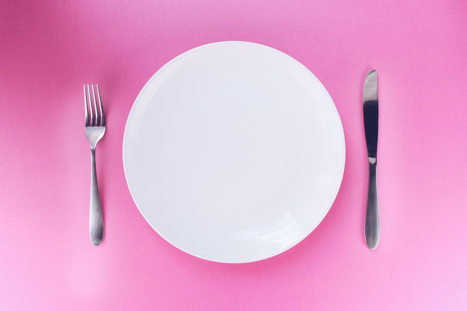 An empty white plate with a fork and knife on a pink background