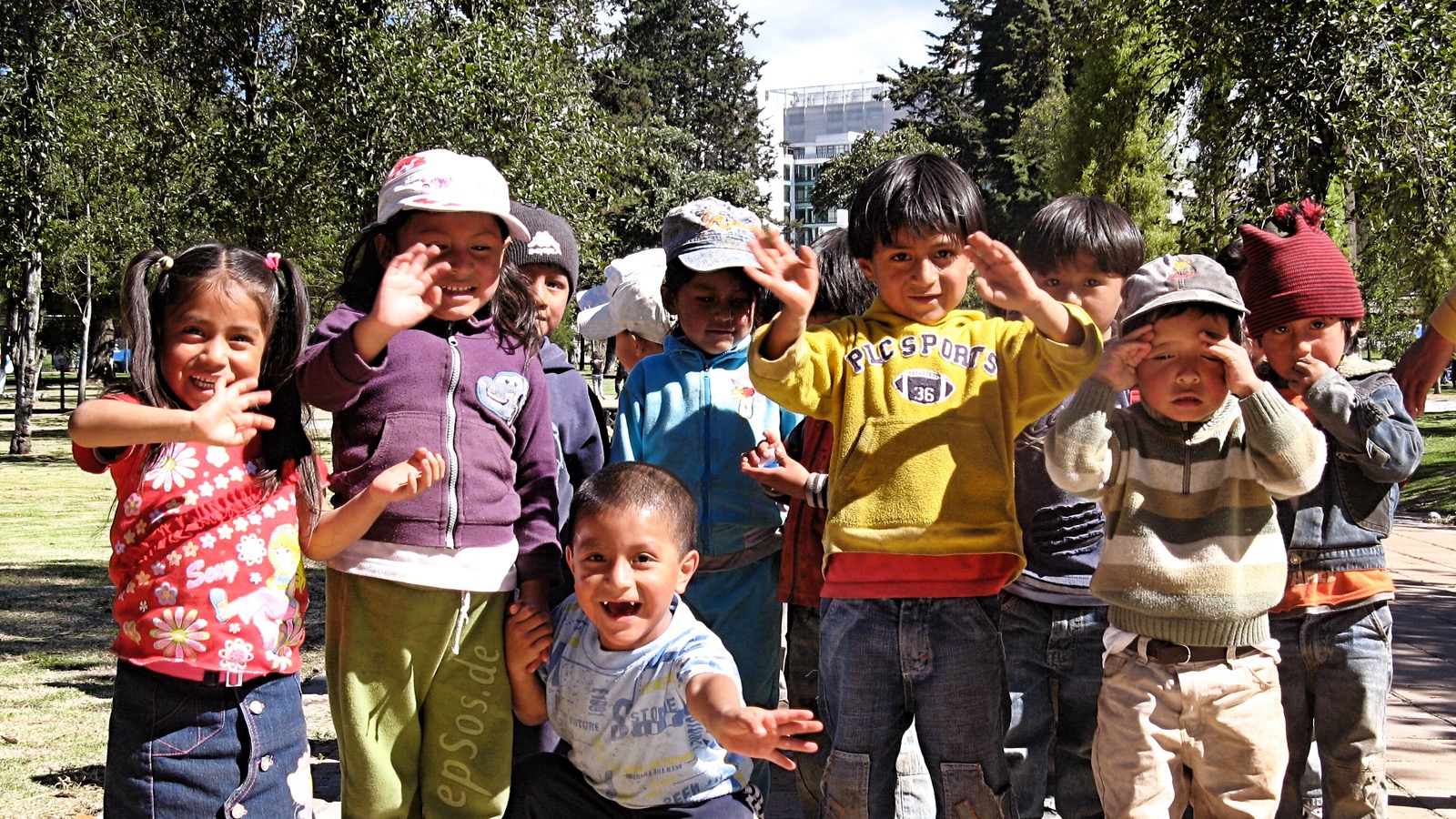 A group of happy, healthy young children smiling and waving outdoors in a park.