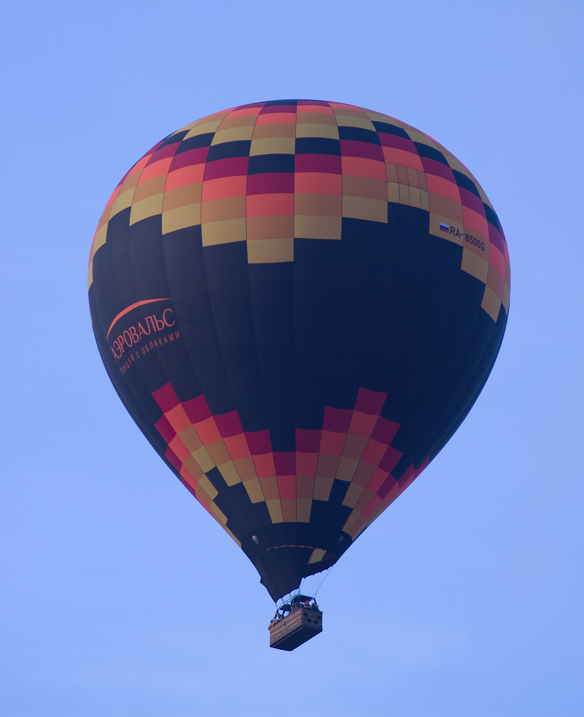 A large hot-air balloon in a checked pattern of black, red and gold floating high in a clear blue sky, with passengers visible in the wicker basket slung underneath