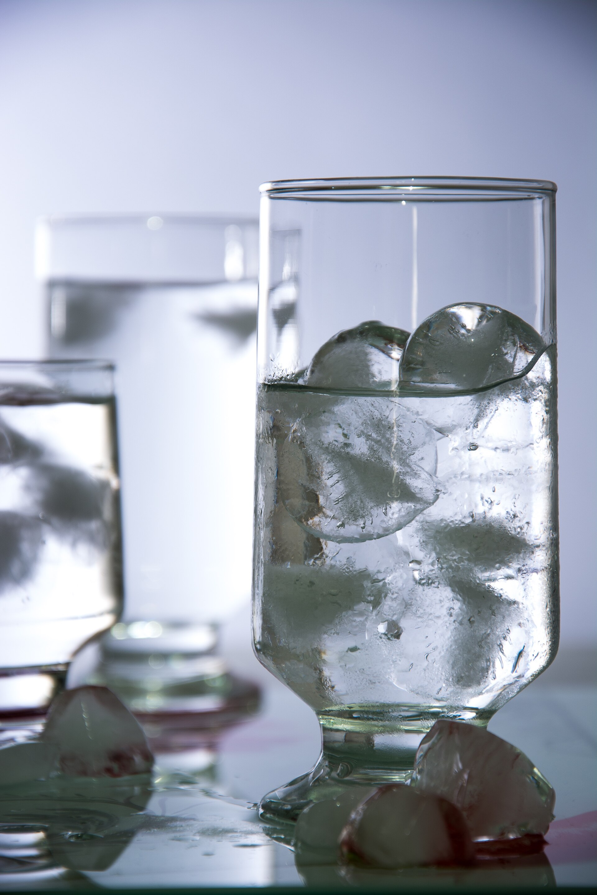A tall glass filled with ice cubes and water, with more ice cubes melting on the surface beside it.