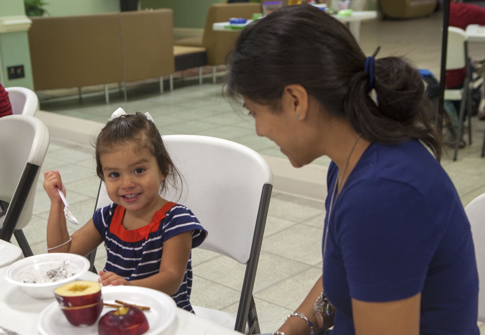 A smiling young child eating a healthy snack, with an apple on the table, at a children's healthy-eating event.