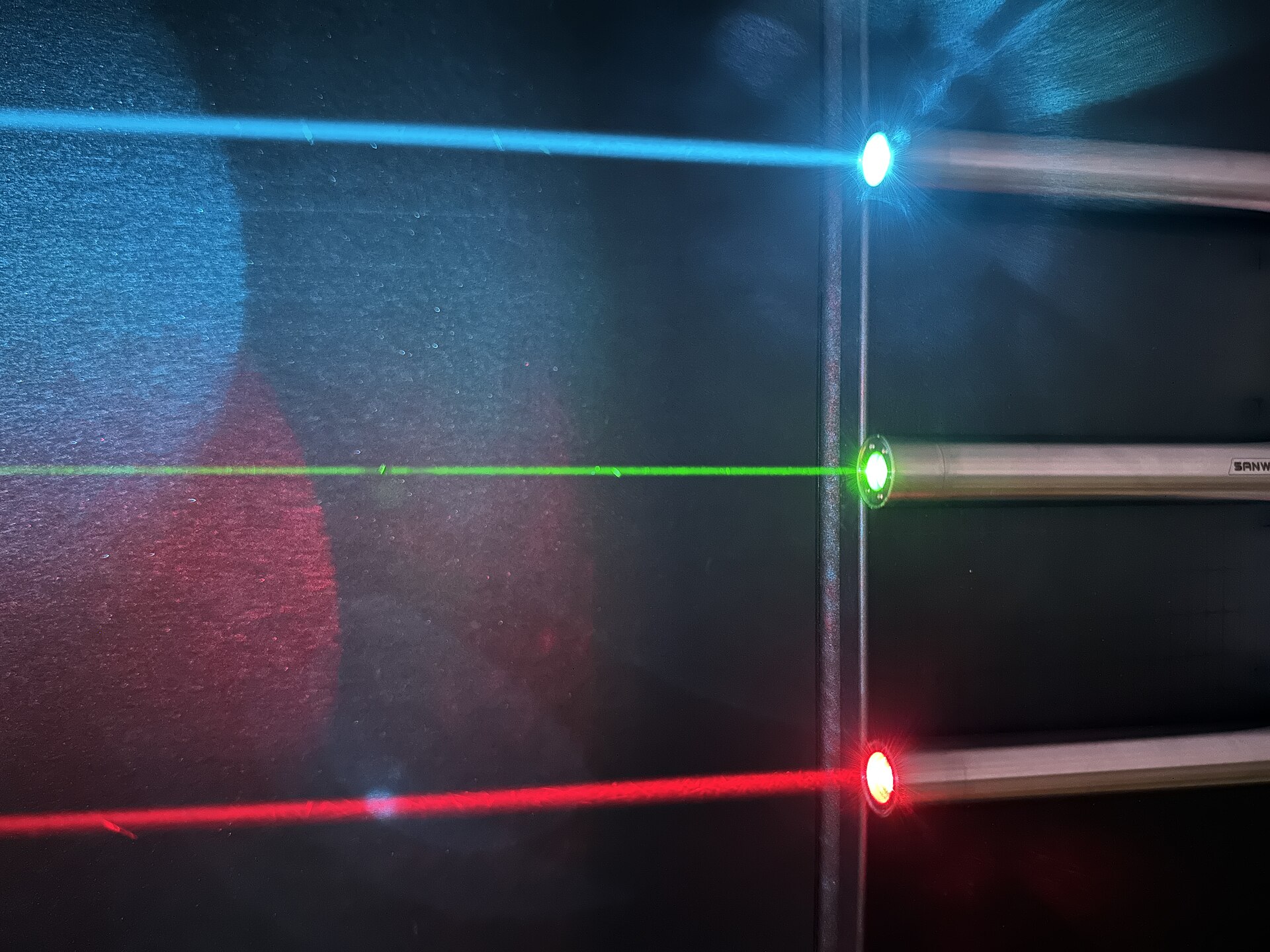 Three laser pointers firing straight, narrow red, green and blue beams across a darkened room