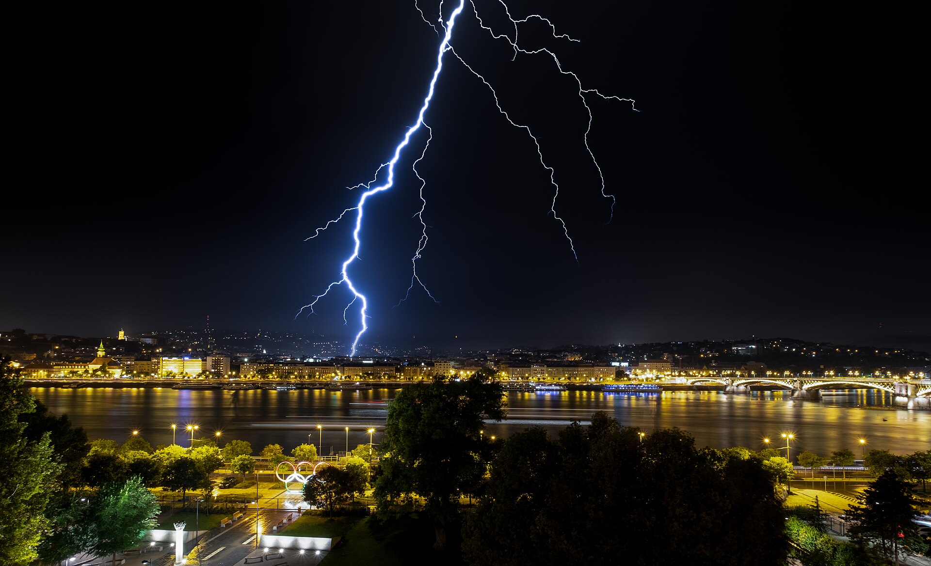 A bright jagged bolt of lightning branching down through a night sky and striking beyond a lit-up city on a riverbank.