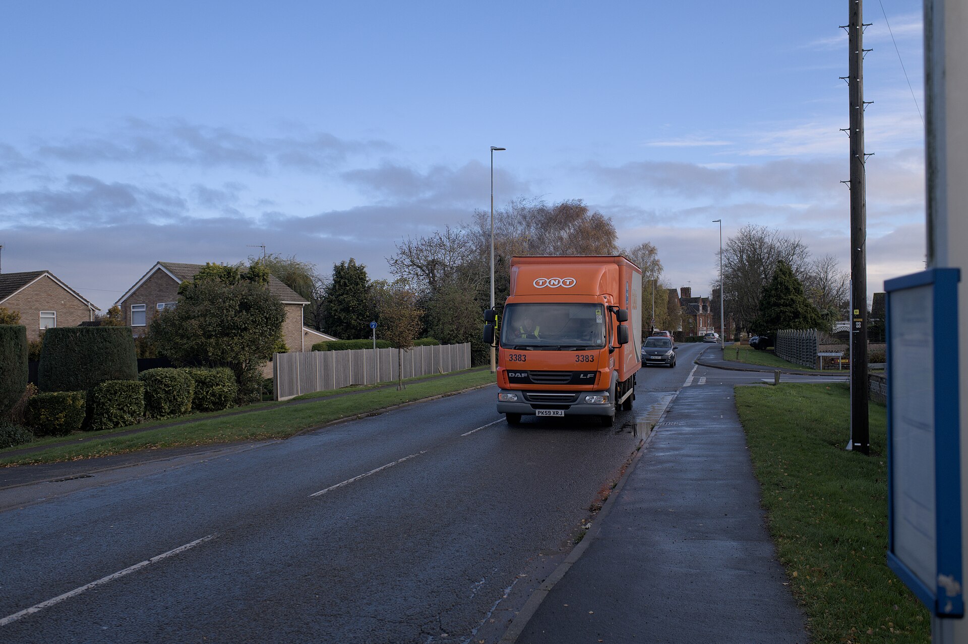 A delivery lorry driving along a residential street past houses on a damp morning.