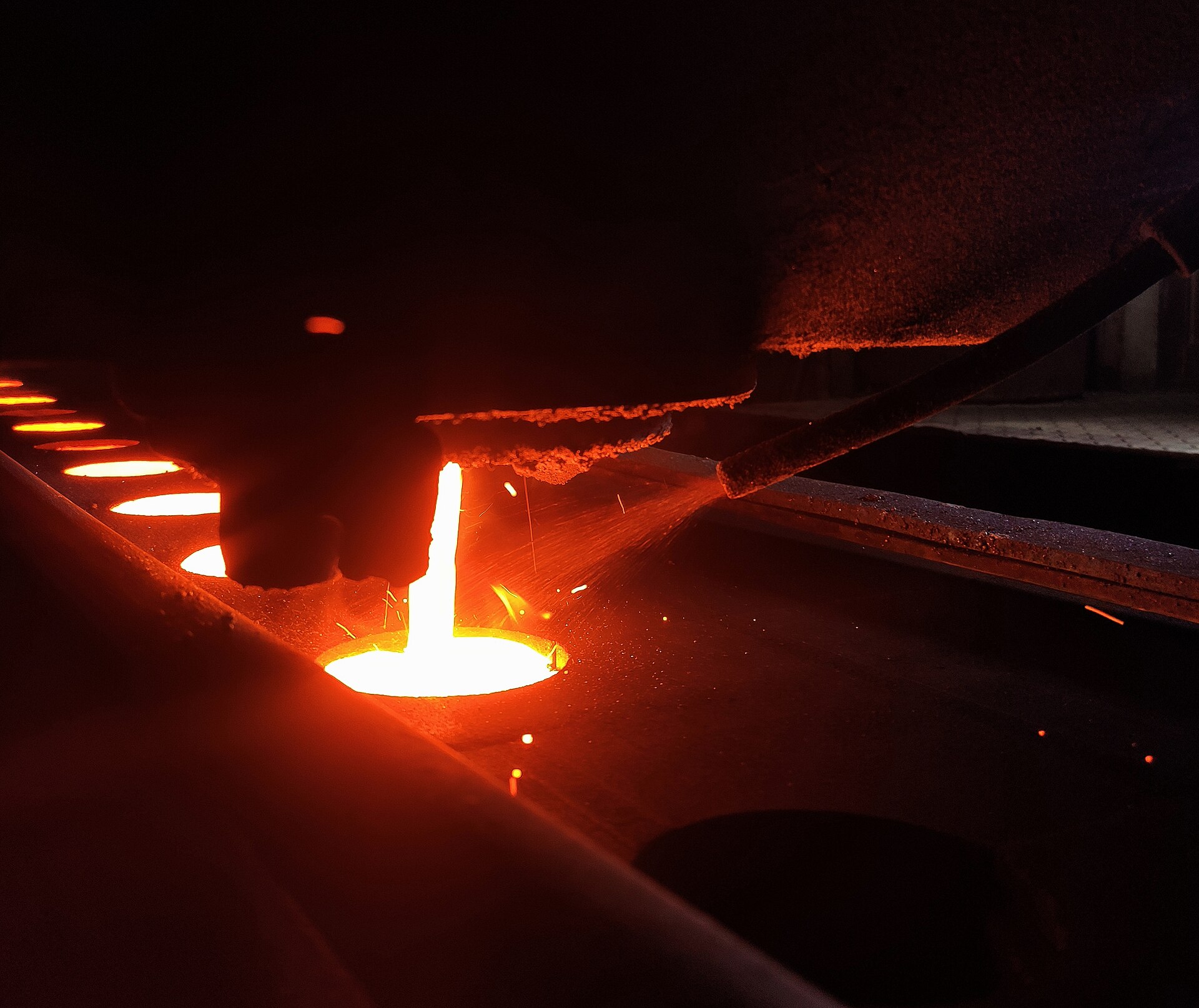 White-hot molten iron pouring in a glowing stream into a mould in a dark foundry, with sparks flying.