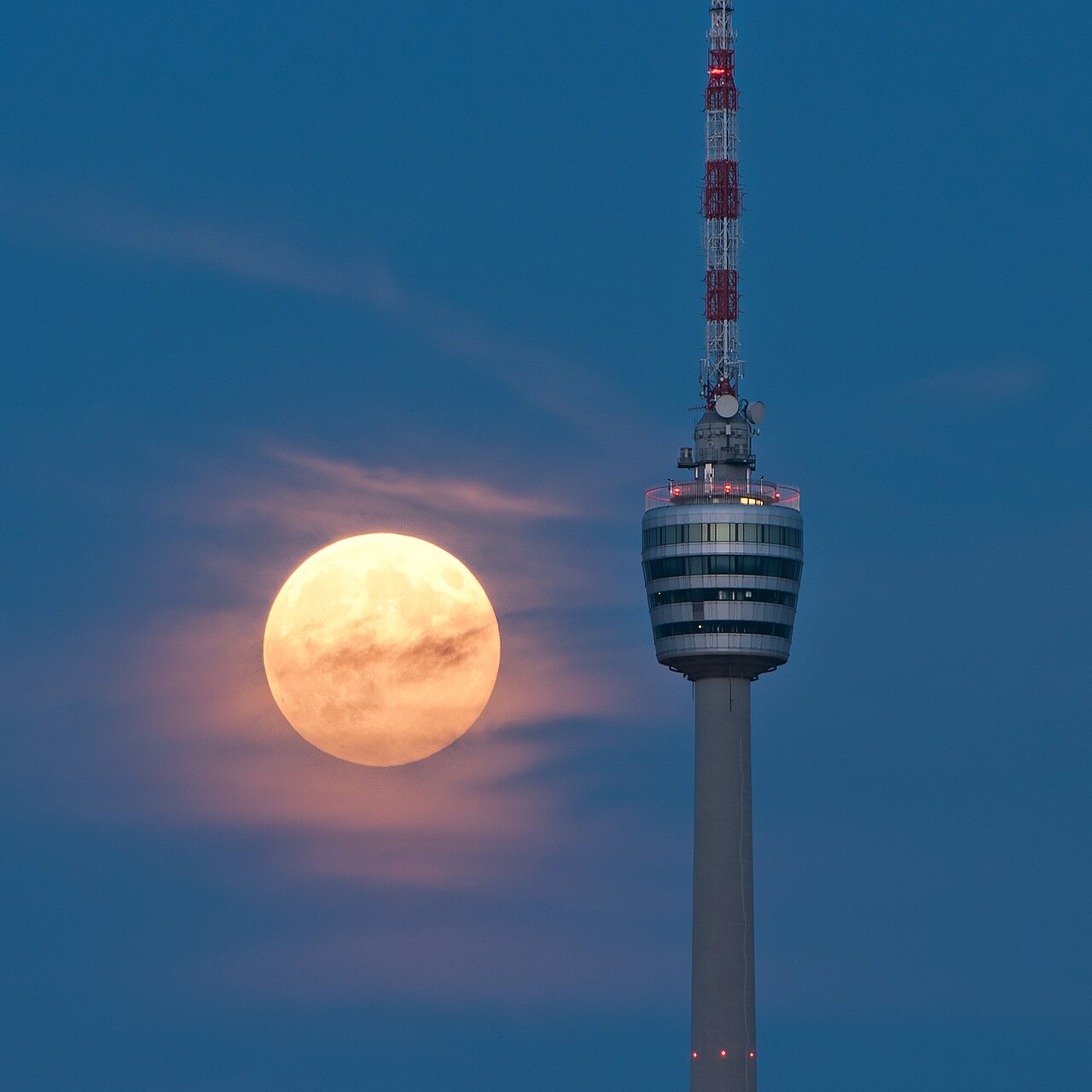 A large orange full Moon low in a deep blue sky, right beside the top of a tall television tower