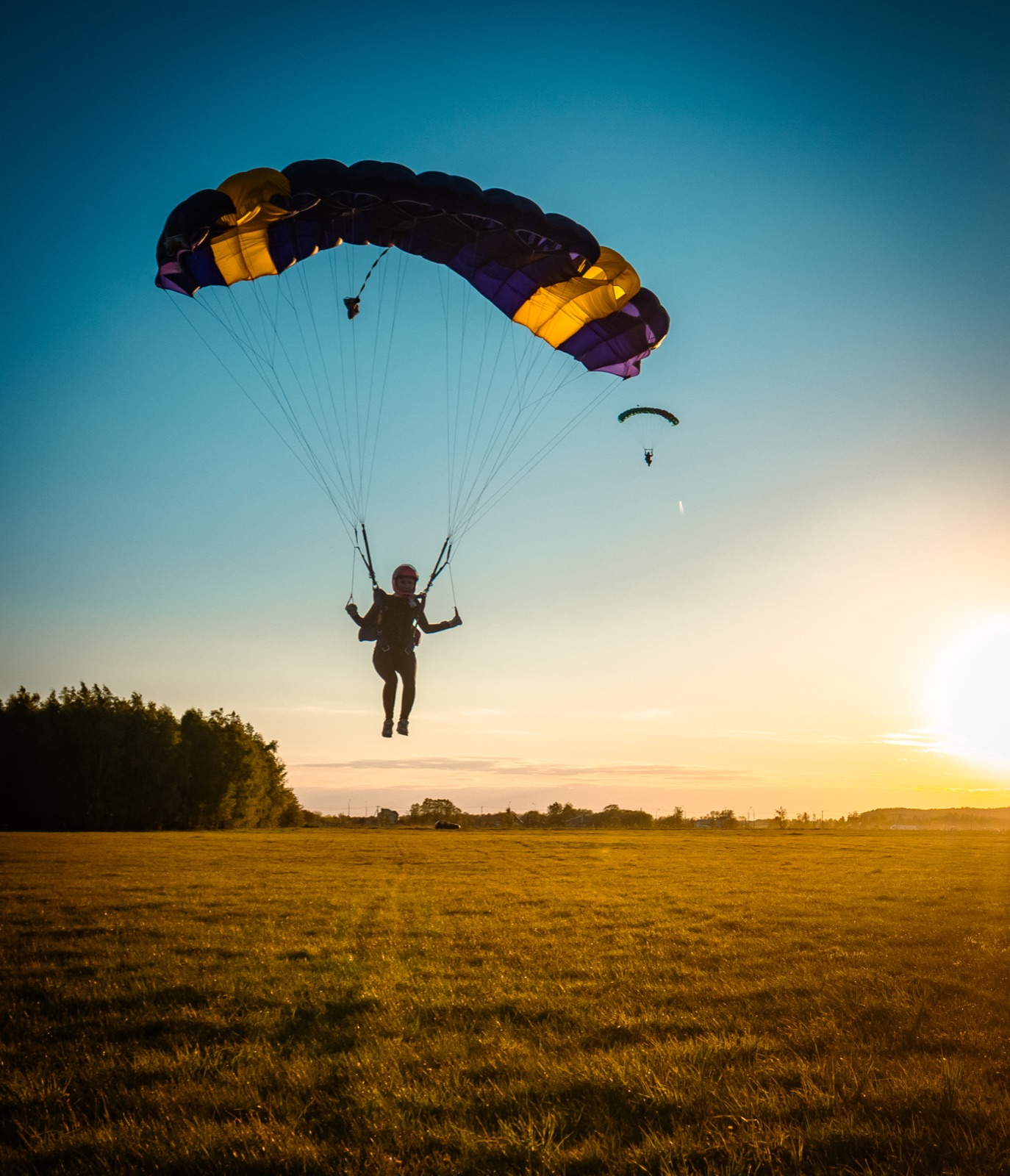 A skydiver descending under a wide open parachute canopy over an open field at sunset