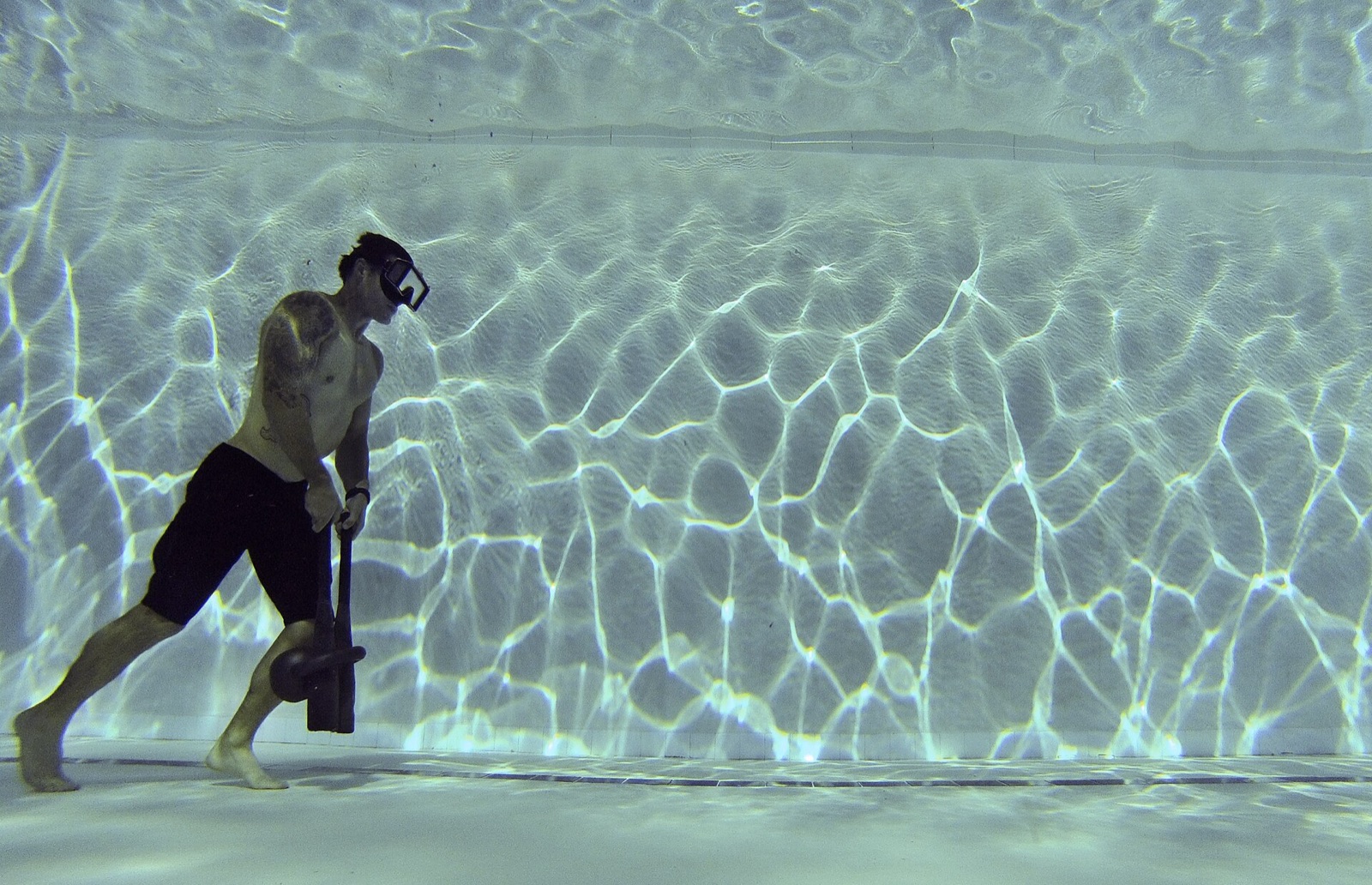 A swimmer in a diving mask walking along the bottom of a deep pool, well below the surface.