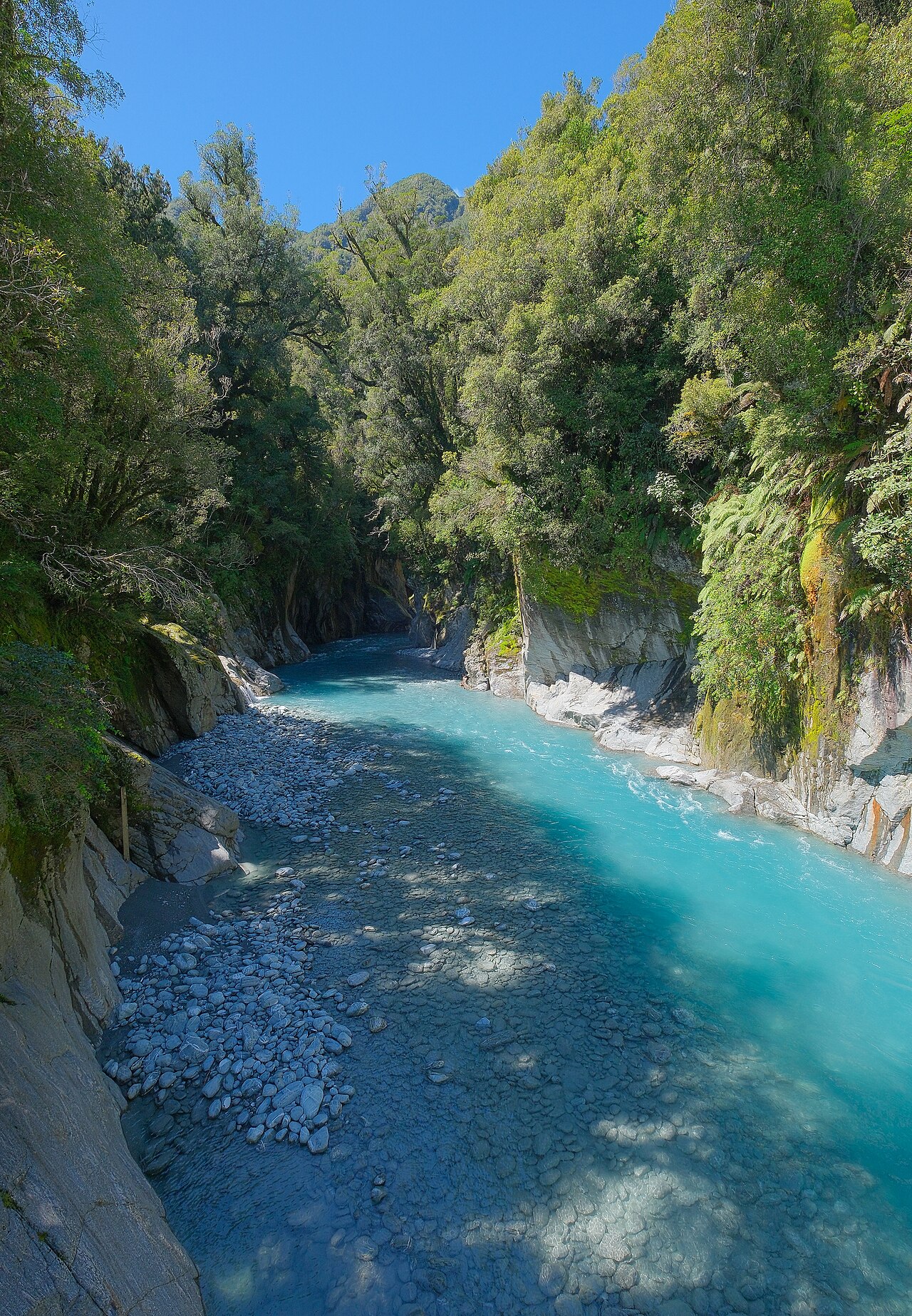 A turquoise river running through a narrow rocky gorge between steep forested walls, with pale cobbles piled along its bed