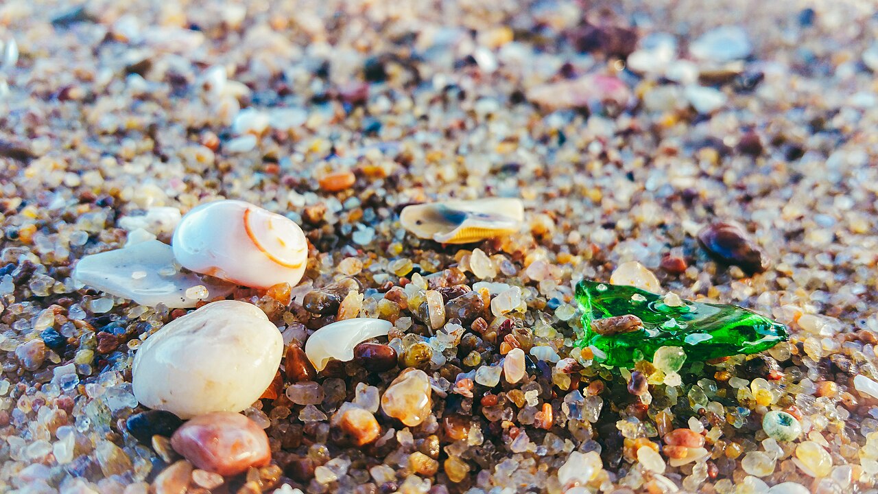 A close-up of beach sand showing thousands of tiny rounded grains mixed with broken shell fragments and a worn piece of green glass