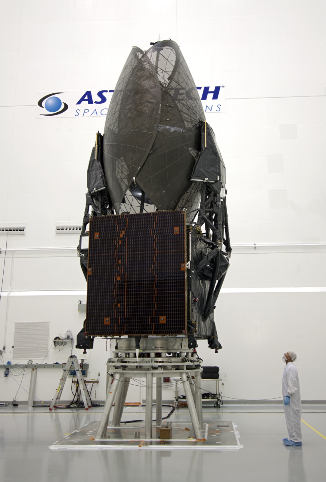 A large satellite being prepared in a clean room before launch, with an engineer beside it for scale.