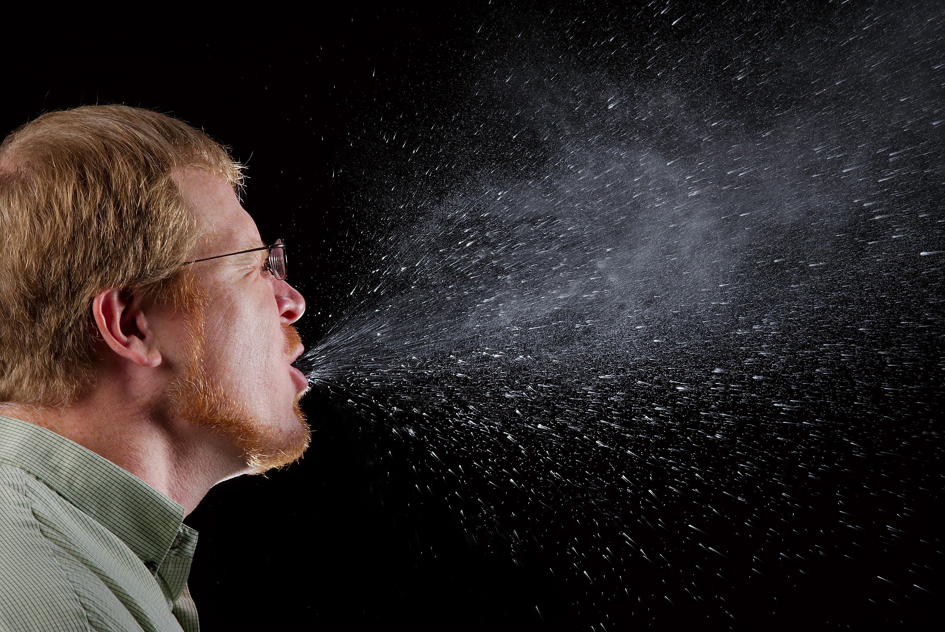 A man caught mid-sneeze against a black background, a huge spray of droplets bursting out and hanging in the air