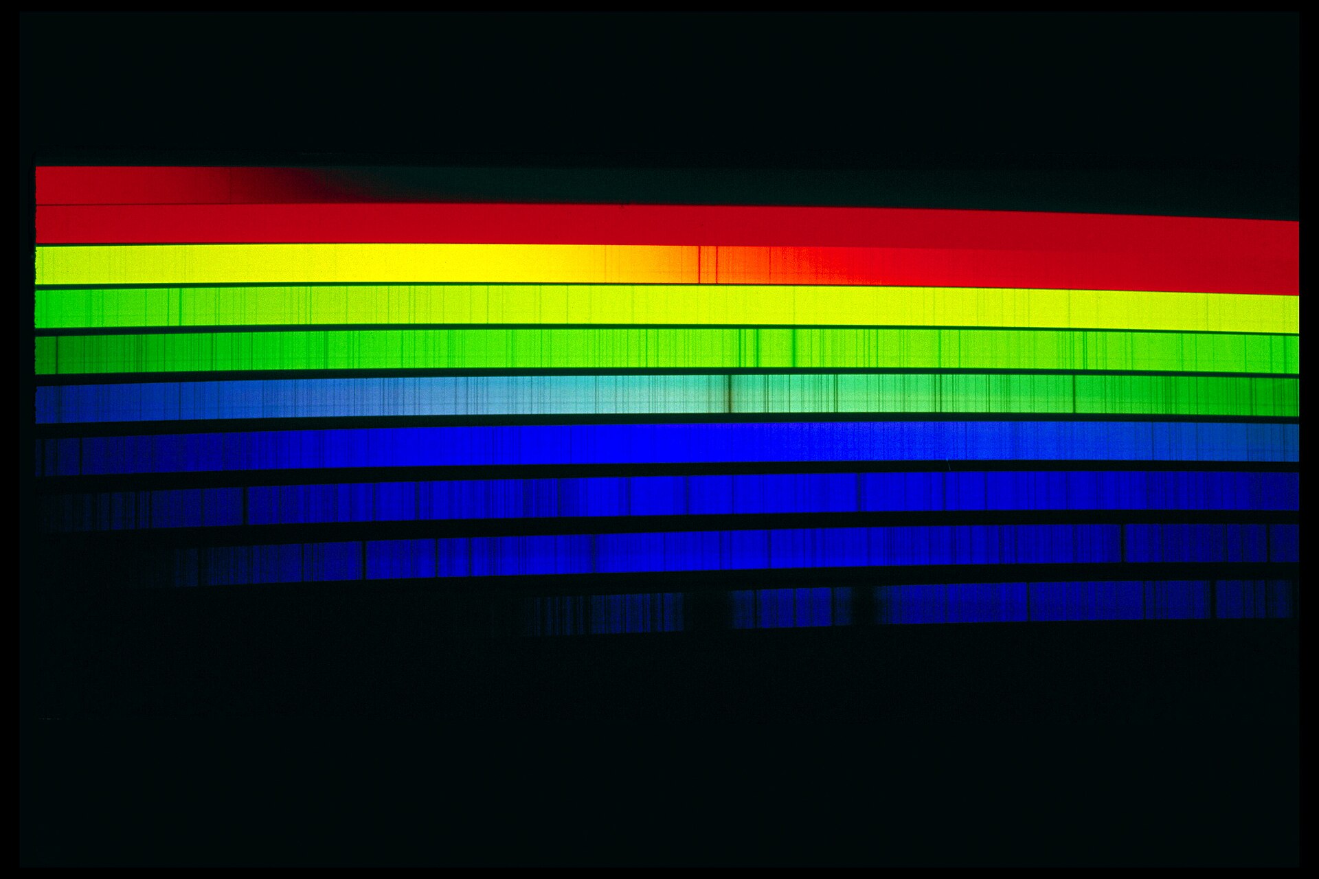 The Sun's spectrum laid out in strips of red, yellow, green and blue, ruled through with hundreds of fine dark lines where elements have swallowed particular colours