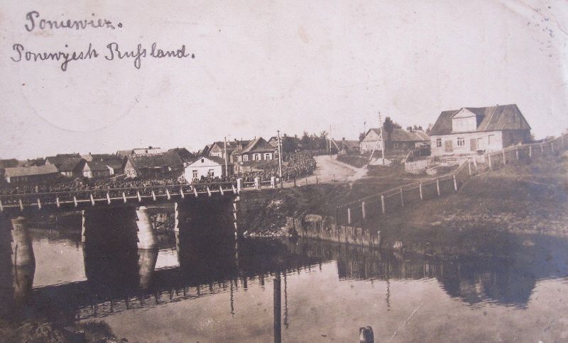 An old photograph of a long column of soldiers marching across a wooden bridge over a river, with a town behind.