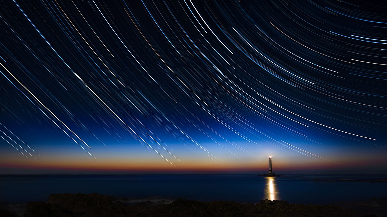 A long exposure of the night sky in which the stars have drawn curved trails, all sweeping around one still point, above a lighthouse at dusk
