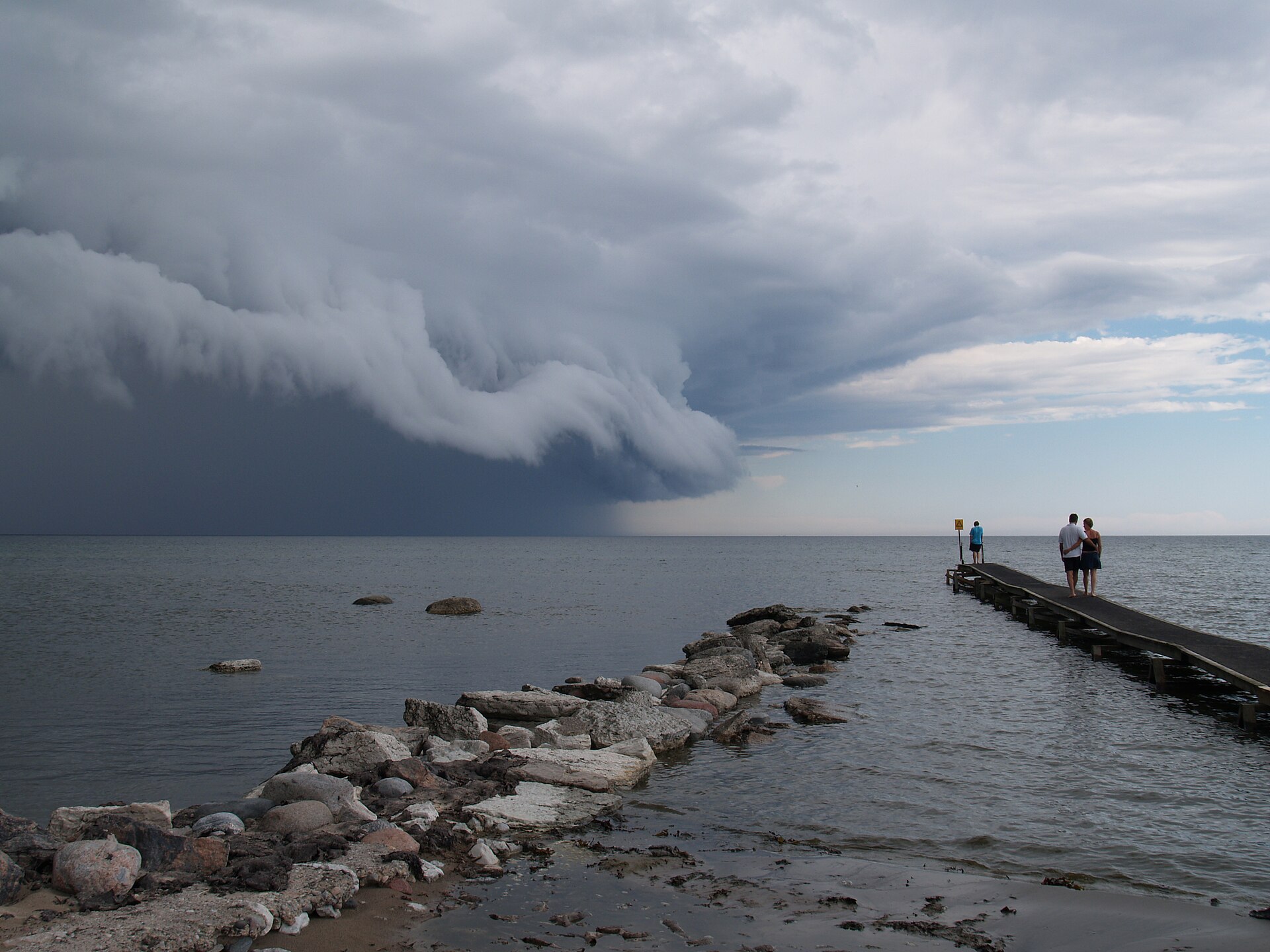 A dark shelf cloud rolling in low over the sea, with rain falling beneath it and people watching from a jetty