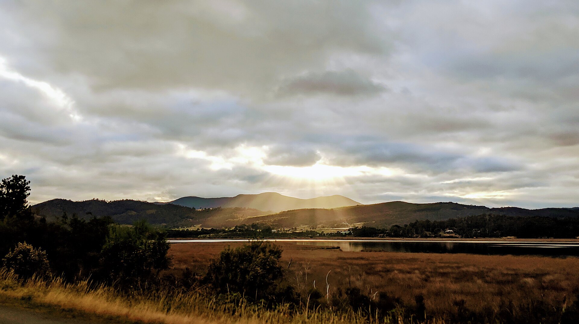 Sunbeams breaking through a gap in heavy cloud and fanning down over hills and water, light arriving at the same speed for everyone who looks