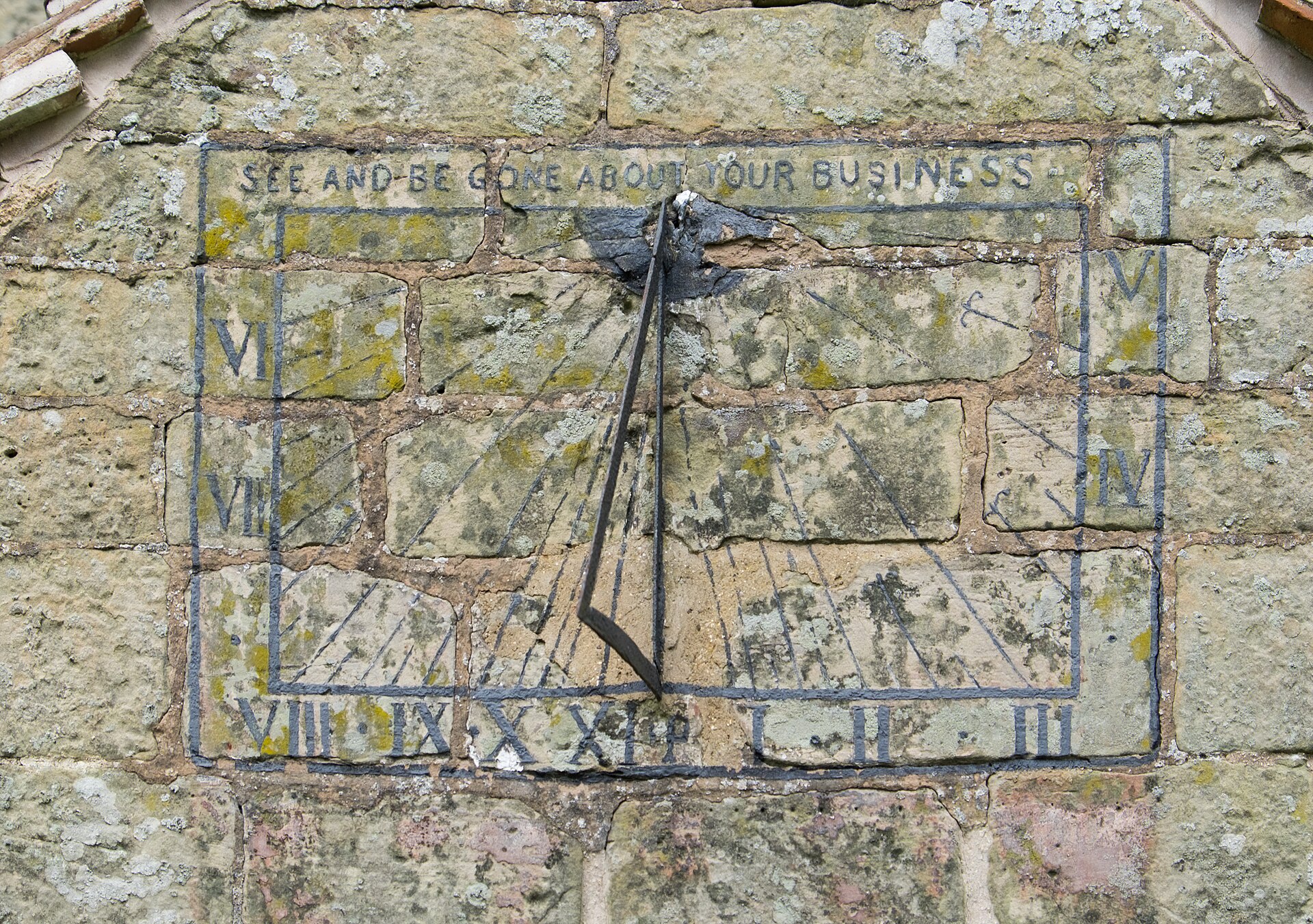 An old stone sundial carved into a lichen-covered church wall, with Roman numerals around the edge, an iron spike casting a shadow, and the motto 'See and be gone about your business'.