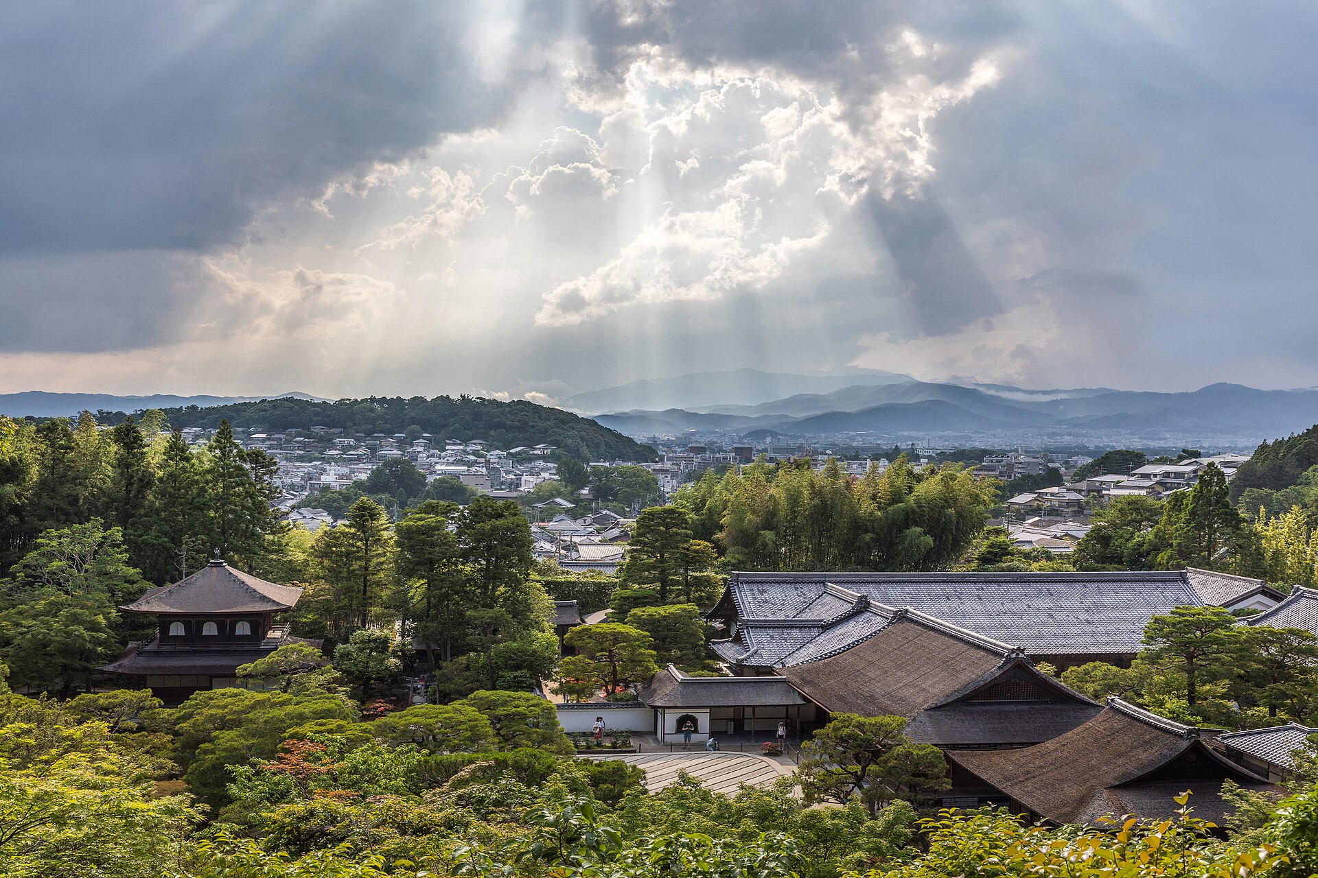 Shafts of sunlight breaking through a gap in heavy cloud and slanting down in straight beams over wooded hills and temple roofs.