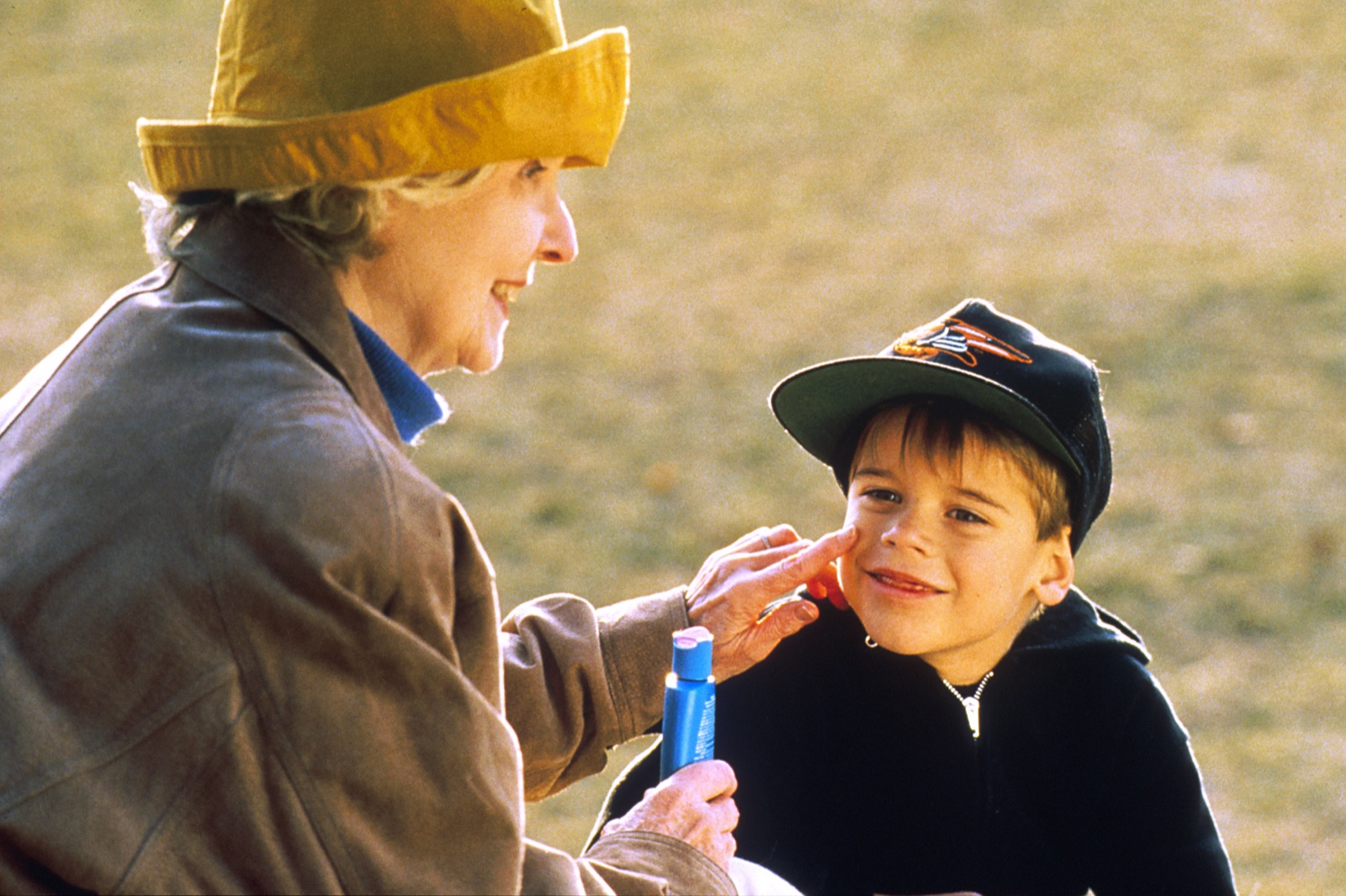 A woman in a sun hat smiling as she dabs sunscreen onto the face of a happy young boy in a cap, outdoors.