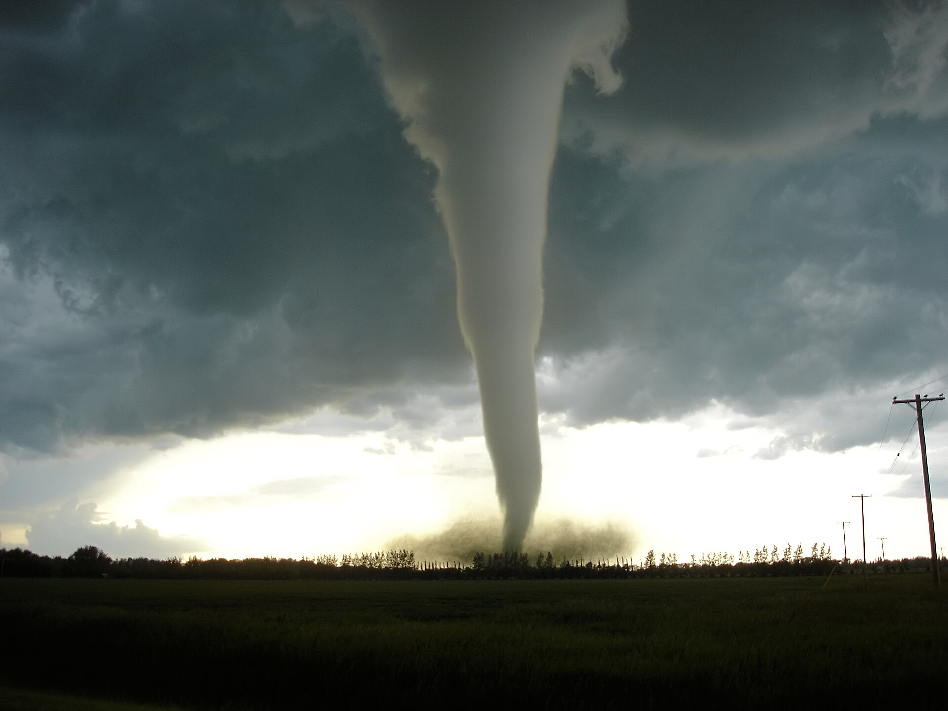 A wide funnel-shaped tornado reaching from a dark storm cloud down to a field, with a cloud of debris swirling where it meets the ground.