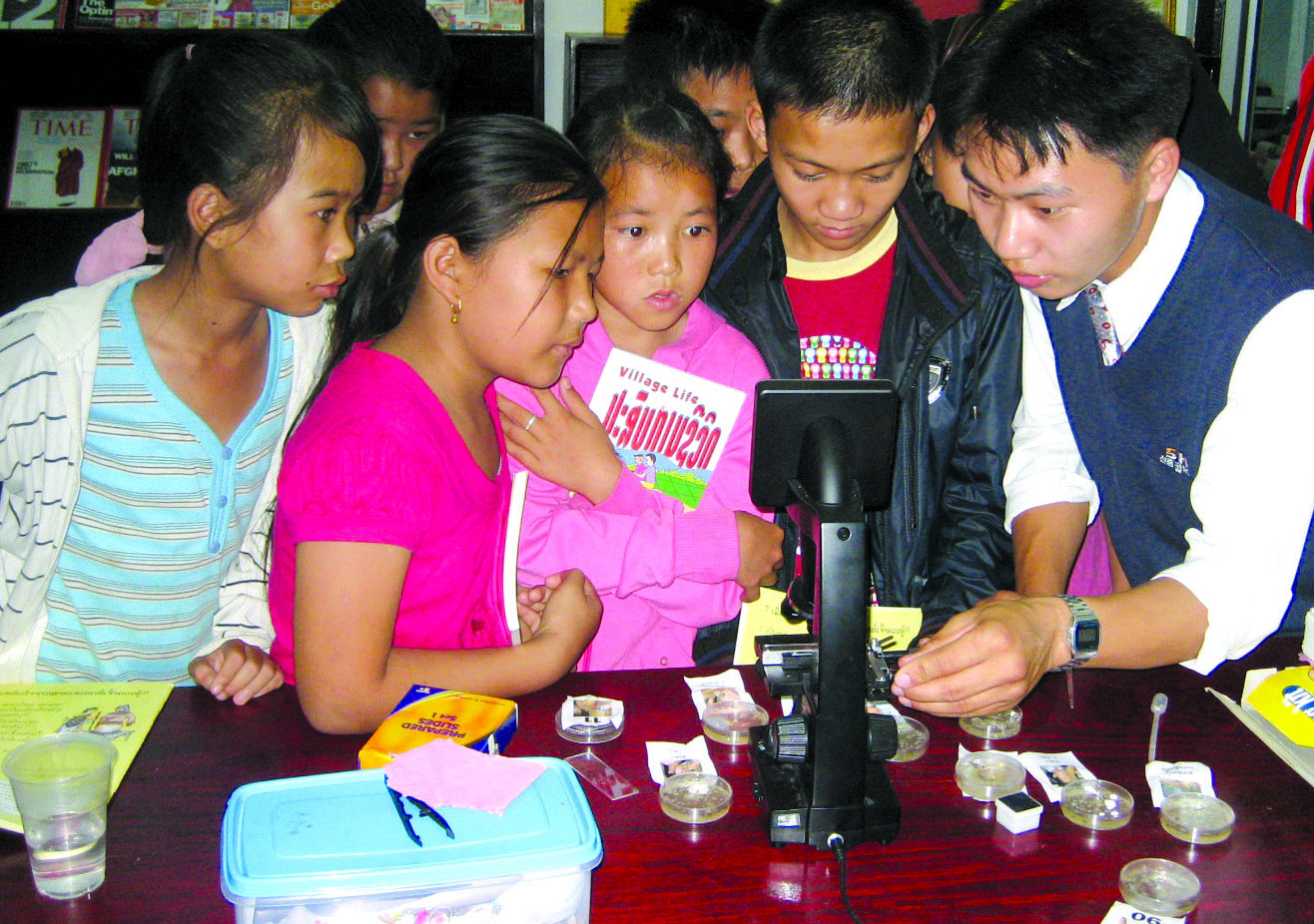 A group of children crowding round a microscope on a table, watching intently as an adult adjusts it.
