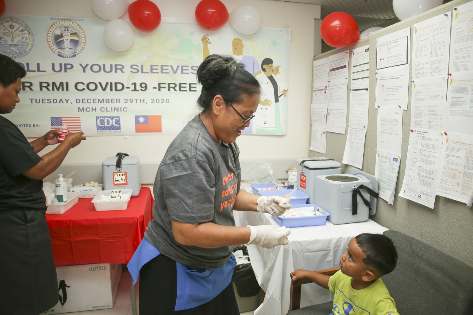 A friendly health worker smiling as she prepares a vaccine at a brightly decorated clinic, with a child looking on.