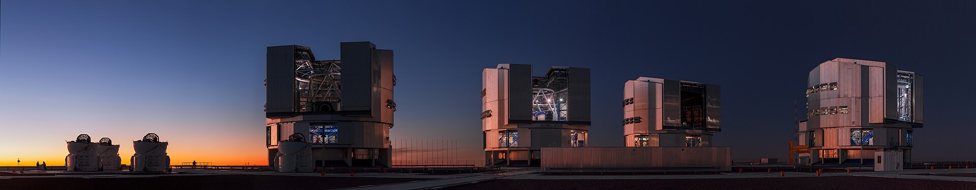 Four huge telescope buildings lined up on a mountain top at twilight, each housing a mirror far wider than any human eye