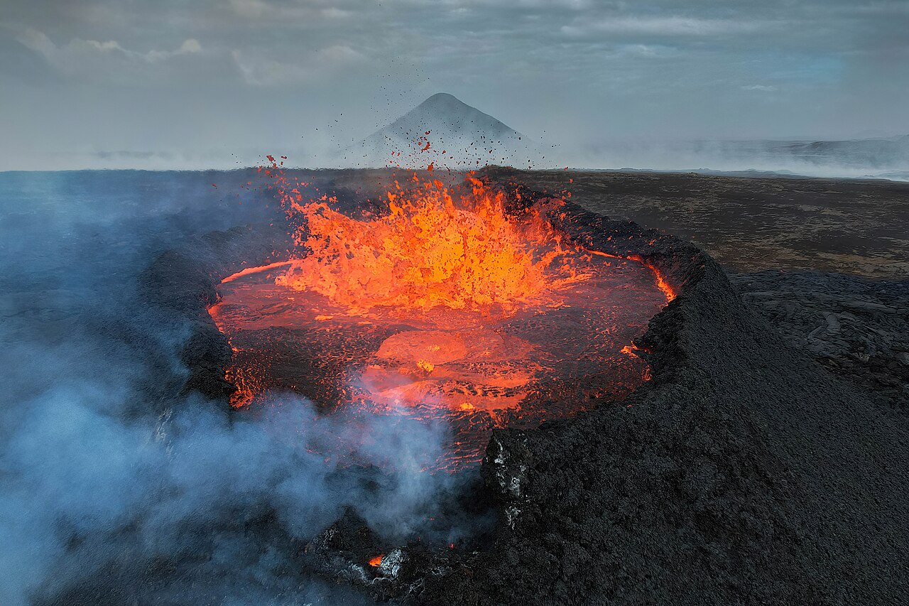 Bright orange molten rock fountaining out of a volcanic crater in Iceland, with steam drifting across the dark lava field